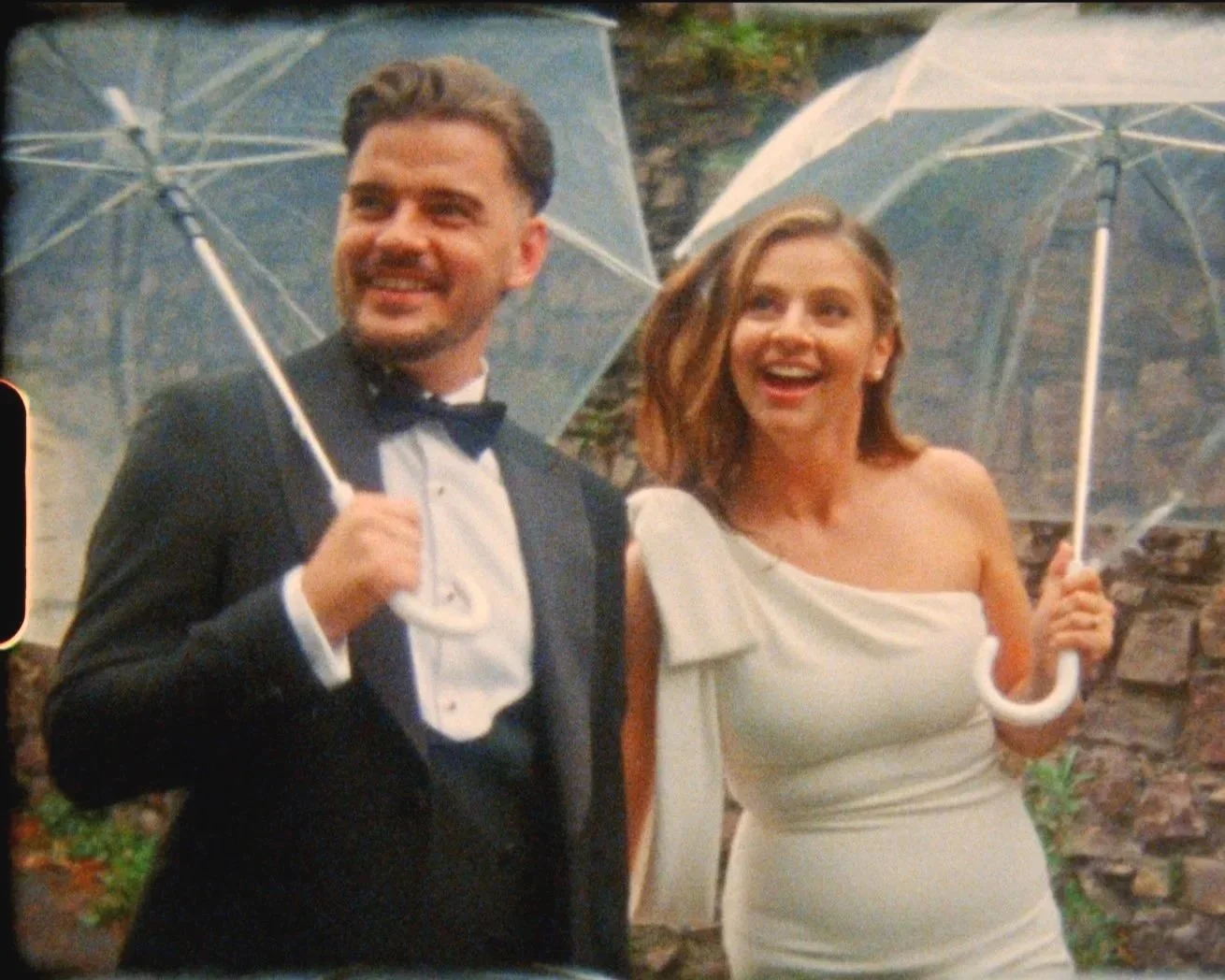 A man and woman dressed in formal attire, holding umbrellas and smiling outdoors.