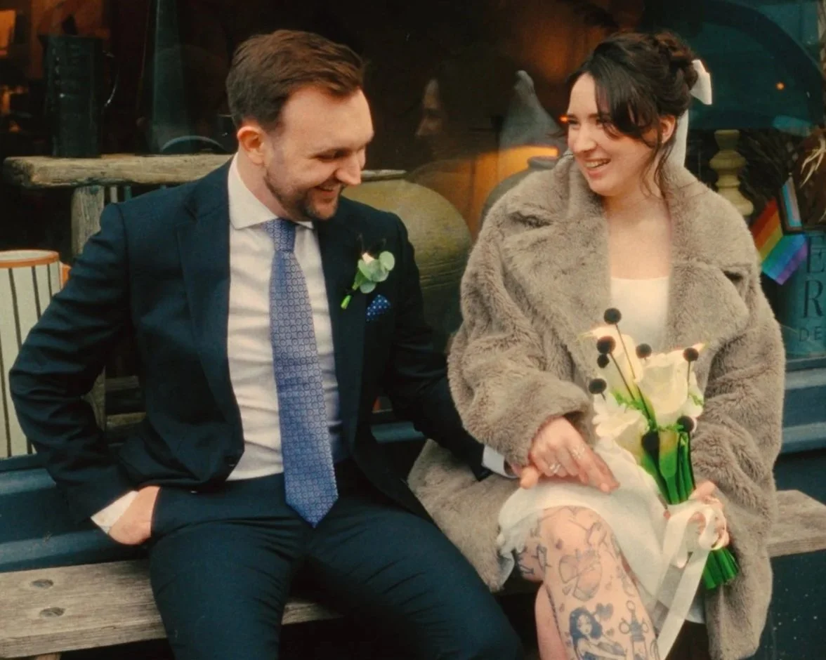 A man in a suit and a woman in a coat sitting on a bench, smiling and holding hands. The woman is holding a bouquet of white flowers. They are in a cozy, outdoor setting with warm lighting.