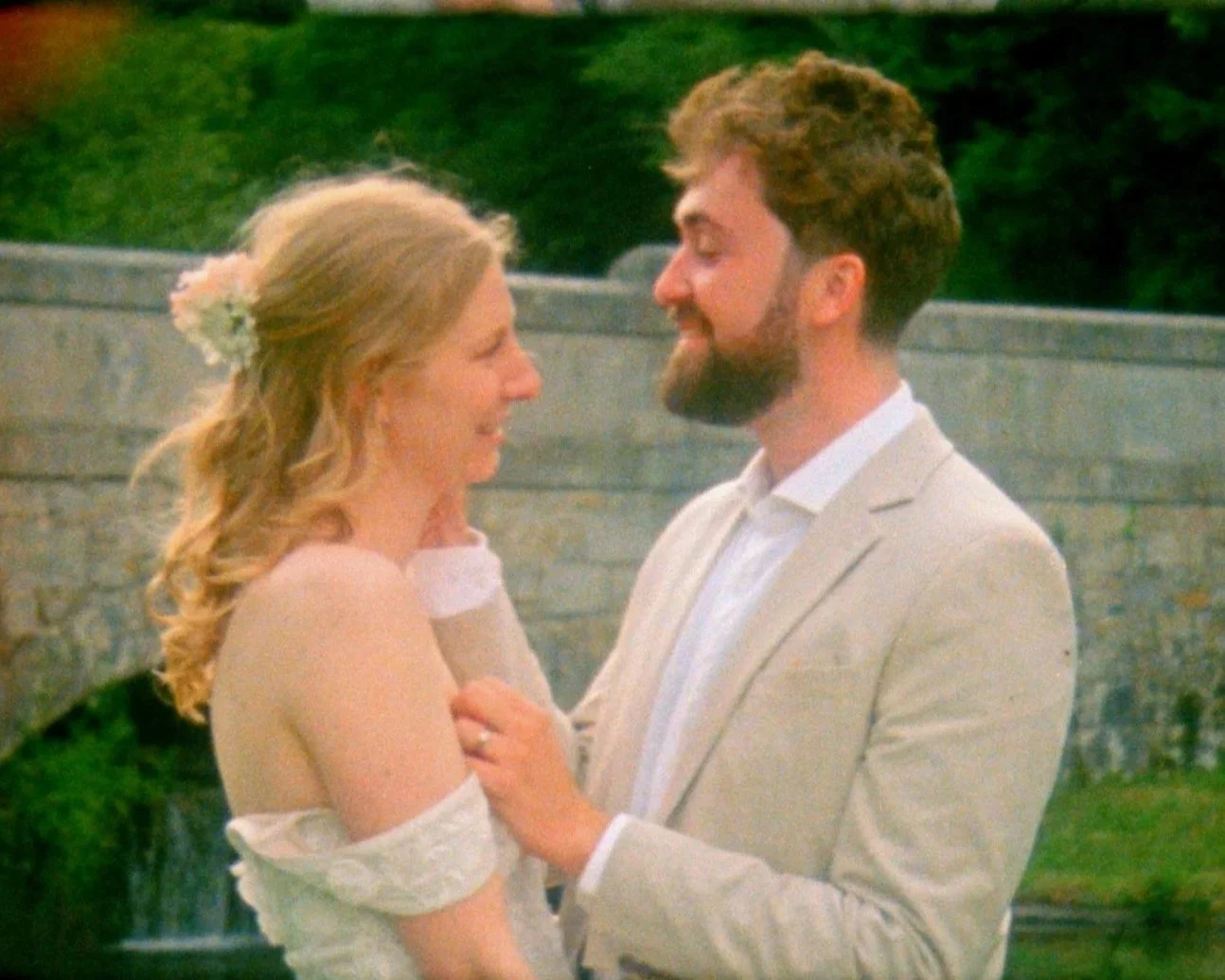 A happy couple on their wedding day, embracing each other outdoors in front of a stone wall and greenery.