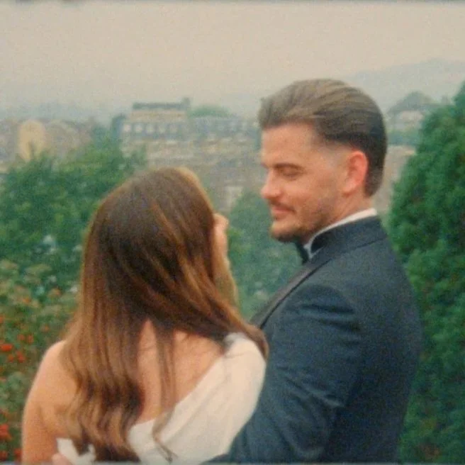 A couple embracing outdoors with greenery and buildings in the background.