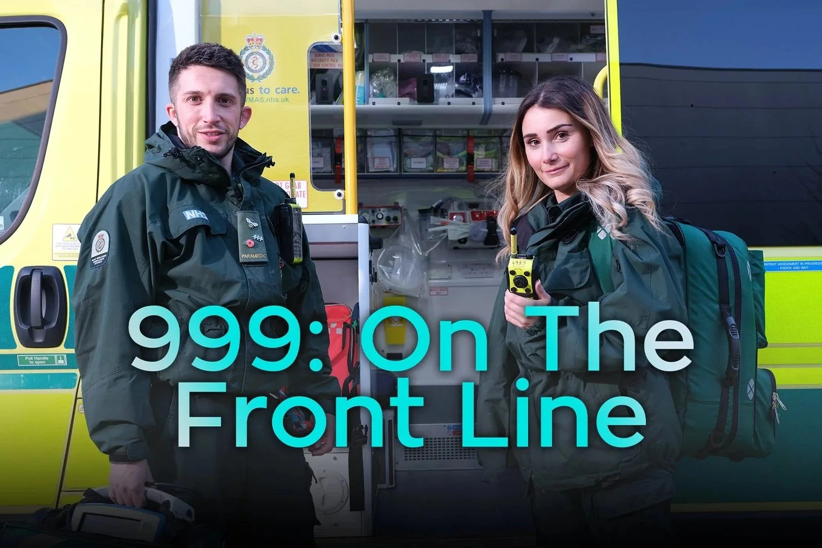 Two emergency medical responders standing in front of an ambulance. The male responder is holding a medical bag and the female responder is holding a radio. They are both wearing dark green uniforms with emergency service patches.