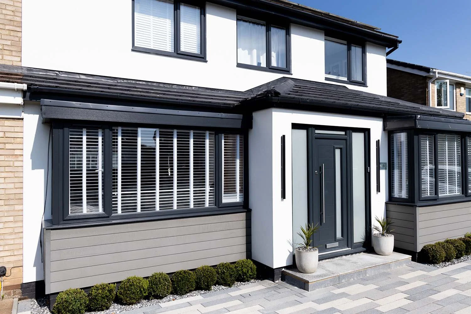 Modern suburban house with white and black exterior, large windows with blinds, and potted plants by the door.