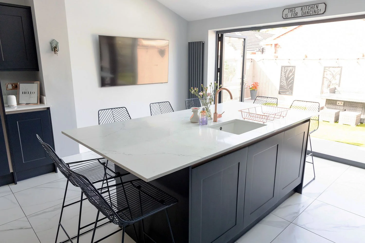 Modern kitchen with a large marble-topped island, black cabinets, and metal bar stools. There's a sink on the island, sliding glass doors to a patio, a wall-mounted TV, and a sign that reads "This Kitchen Is For Dancing!"