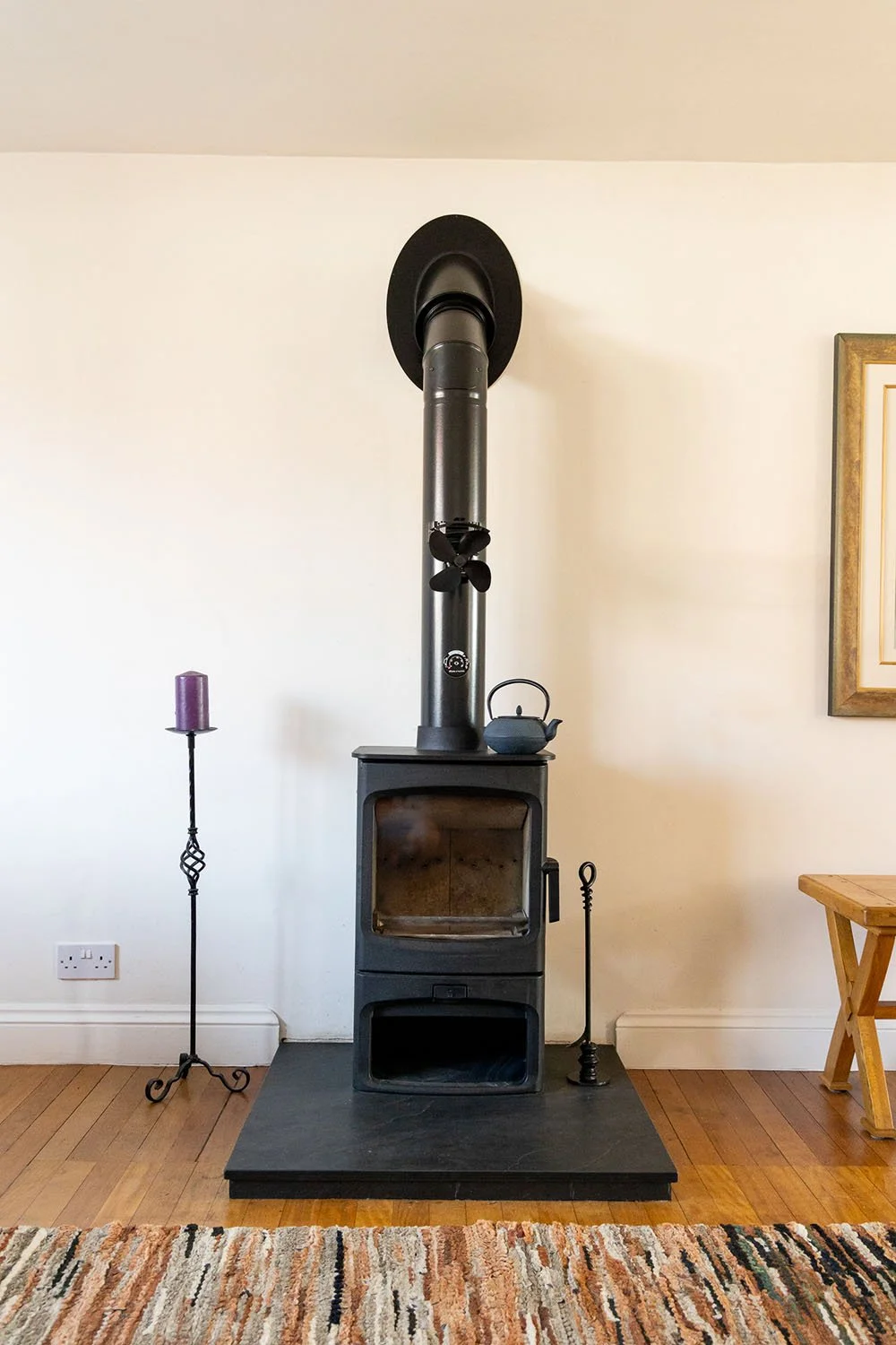 Wood-burning stove with chimney, purple candle on stand, and kettle, set on a wooden floor with a patterned rug."}