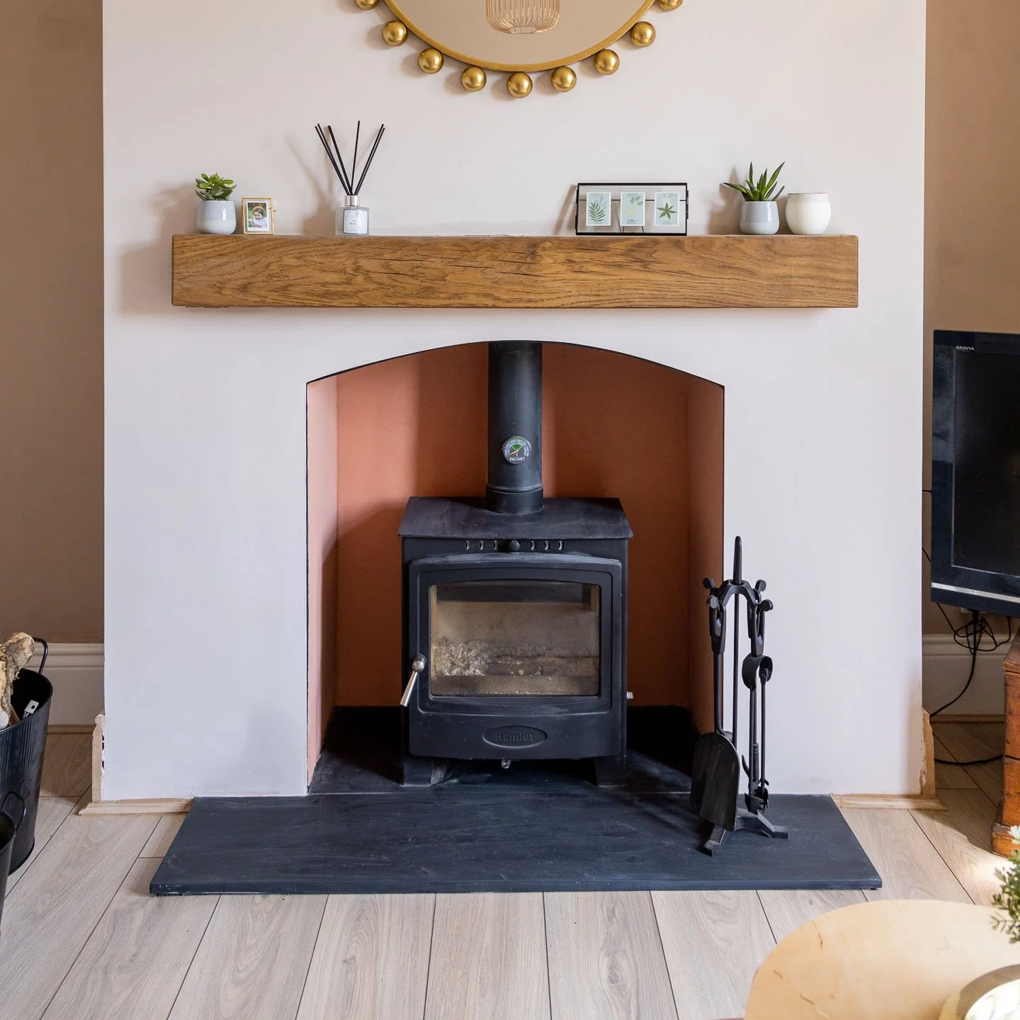A cozy indoor scene with a wood-burning stove placed in a hearth. The stove has a glass front and a black chimney. Above it, a wooden mantel holds small plants, framed art, and a candle. A round mirror with a gold decorative edge hangs on the wall. A set of fireplace tools is nearby on the dark hearthstone, and light wooden flooring is visible.