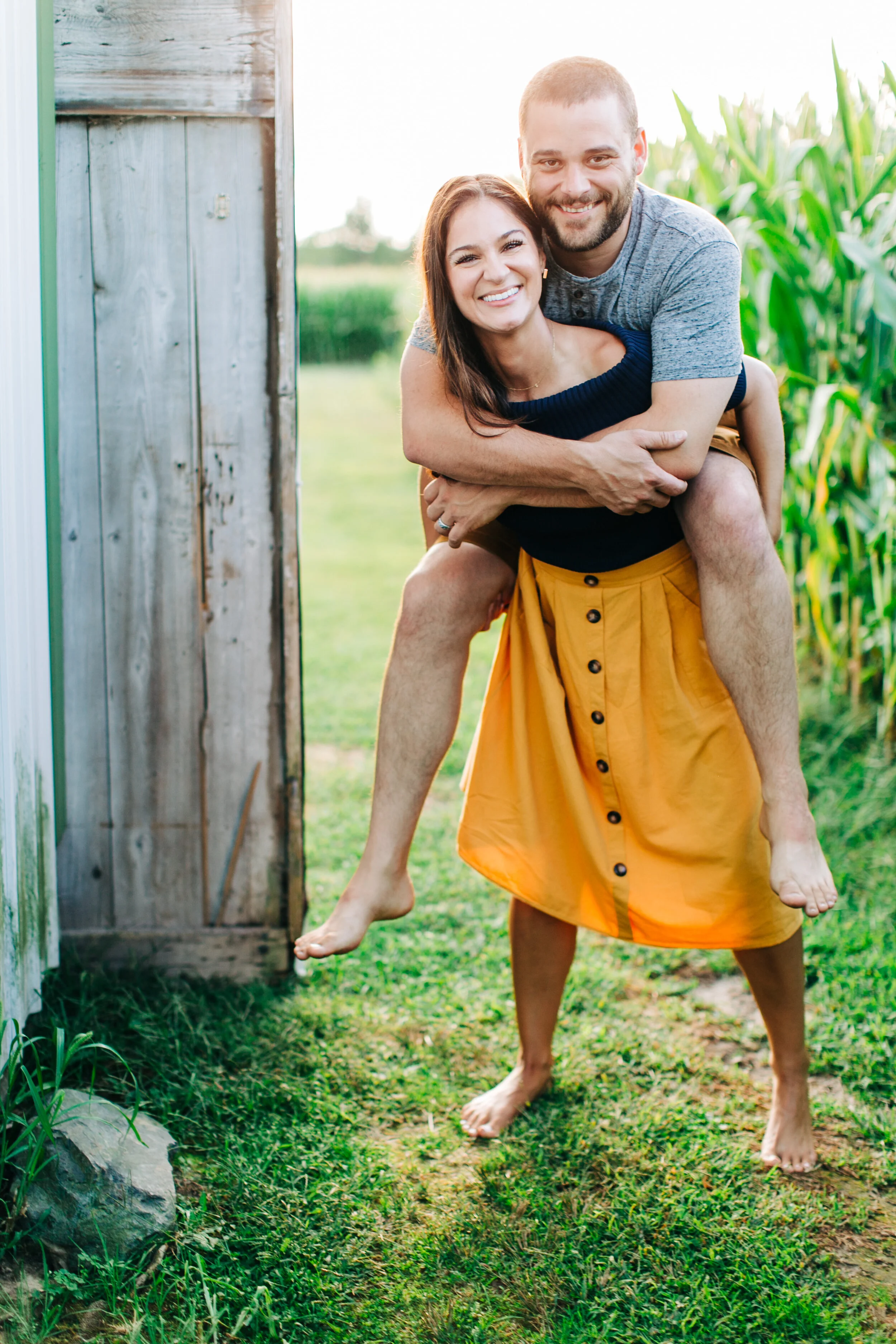 A woman gives her partner a piggyback ride while they smile at the camera, standing barefoot in a grassy garden beside a wooden gate and tall corn stalks.