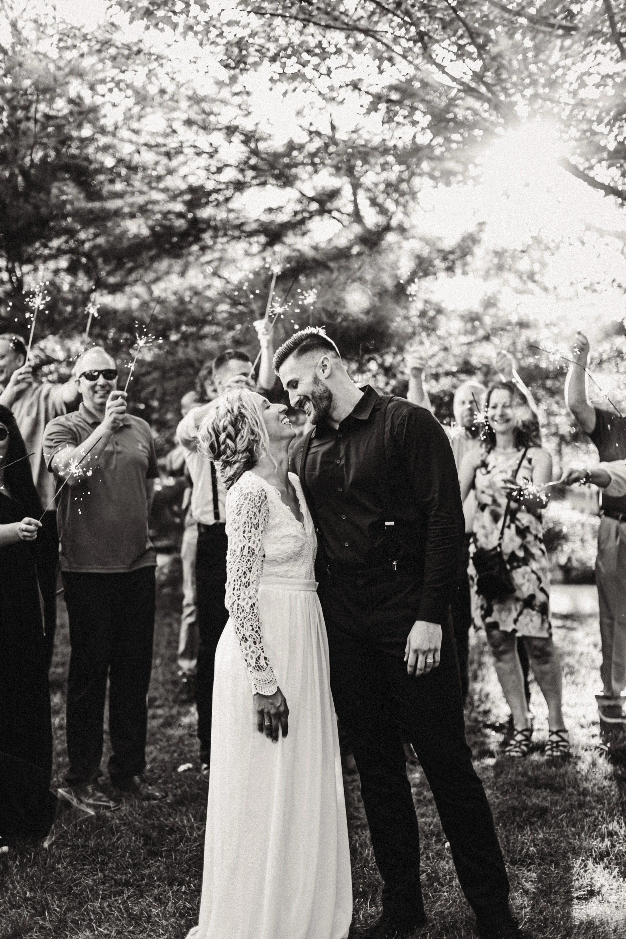 A bride in a long-sleeved lace wedding gown and a groom in a dark dress shirt share a quiet, intimate moment while guests surround them holding sparklers.