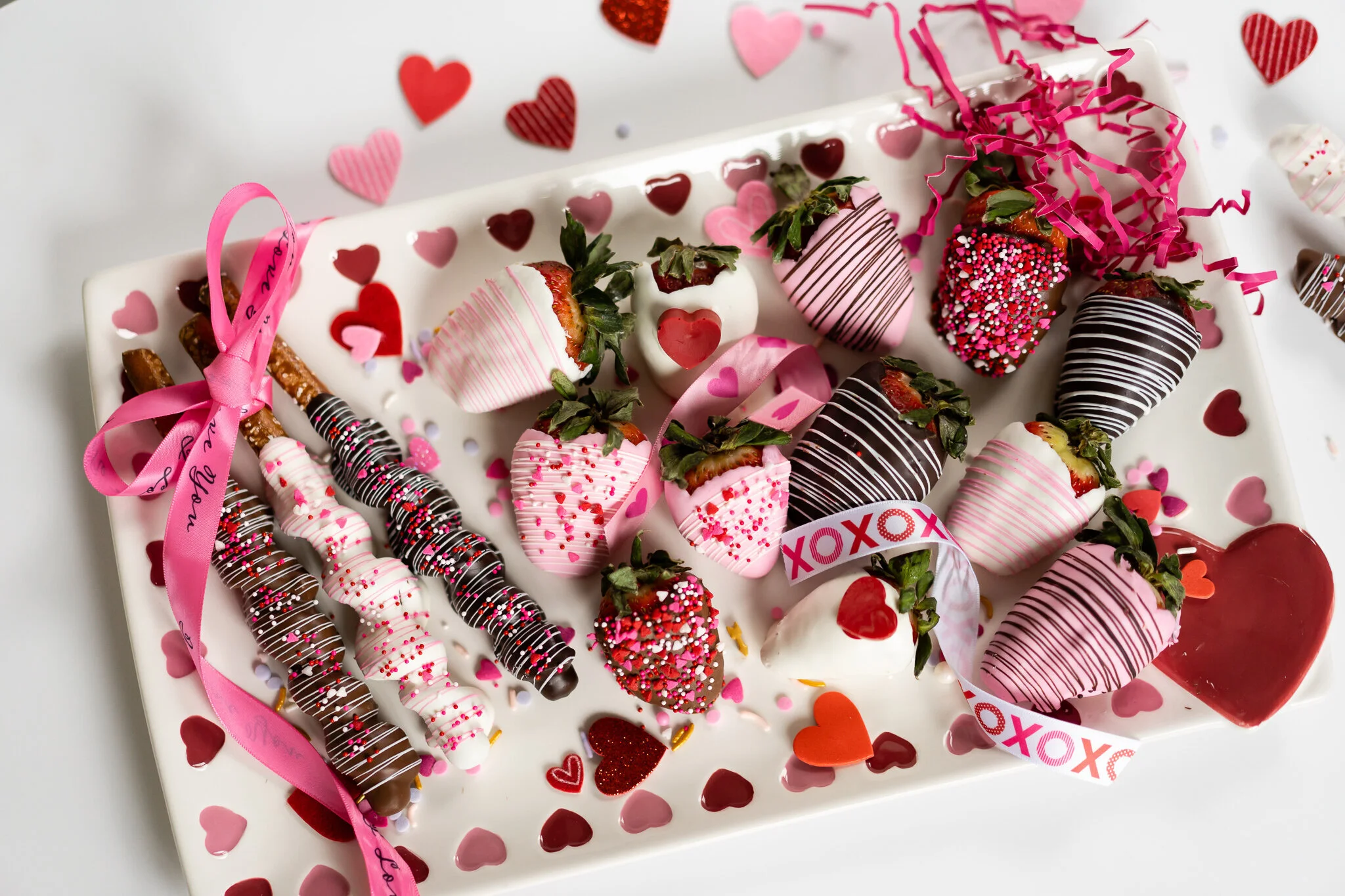 A flat-lay photo of assorted chocolate-covered strawberries and decorated pretzel rods arranged on a white tray with red and pink heart cutouts, surrounded by Valentine confetti and ribbon.