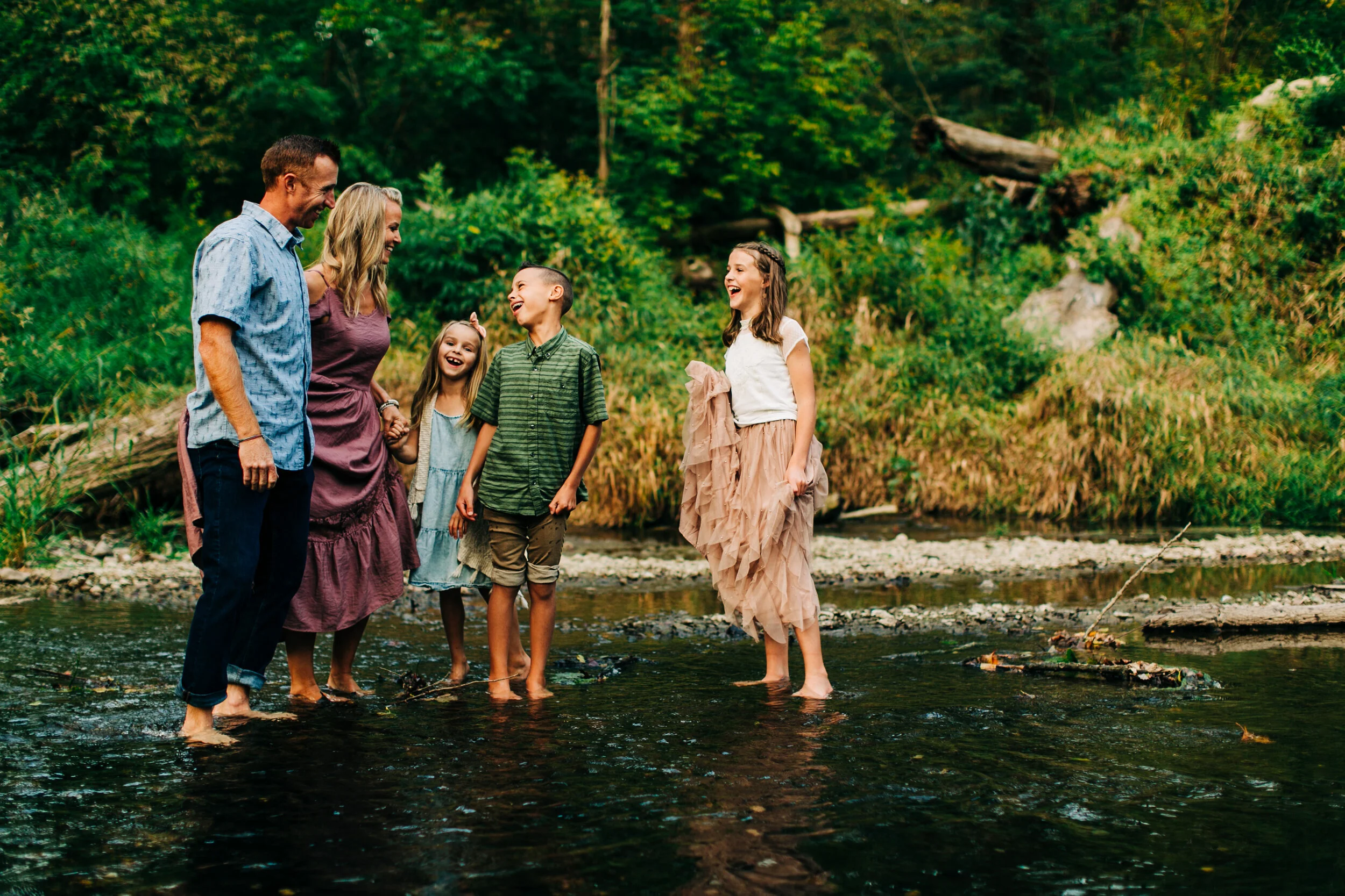 A joyful family of five playfully standing together in a river, barefoot, with green trees and nature surrounding them. Everyone is smiling and having fun.