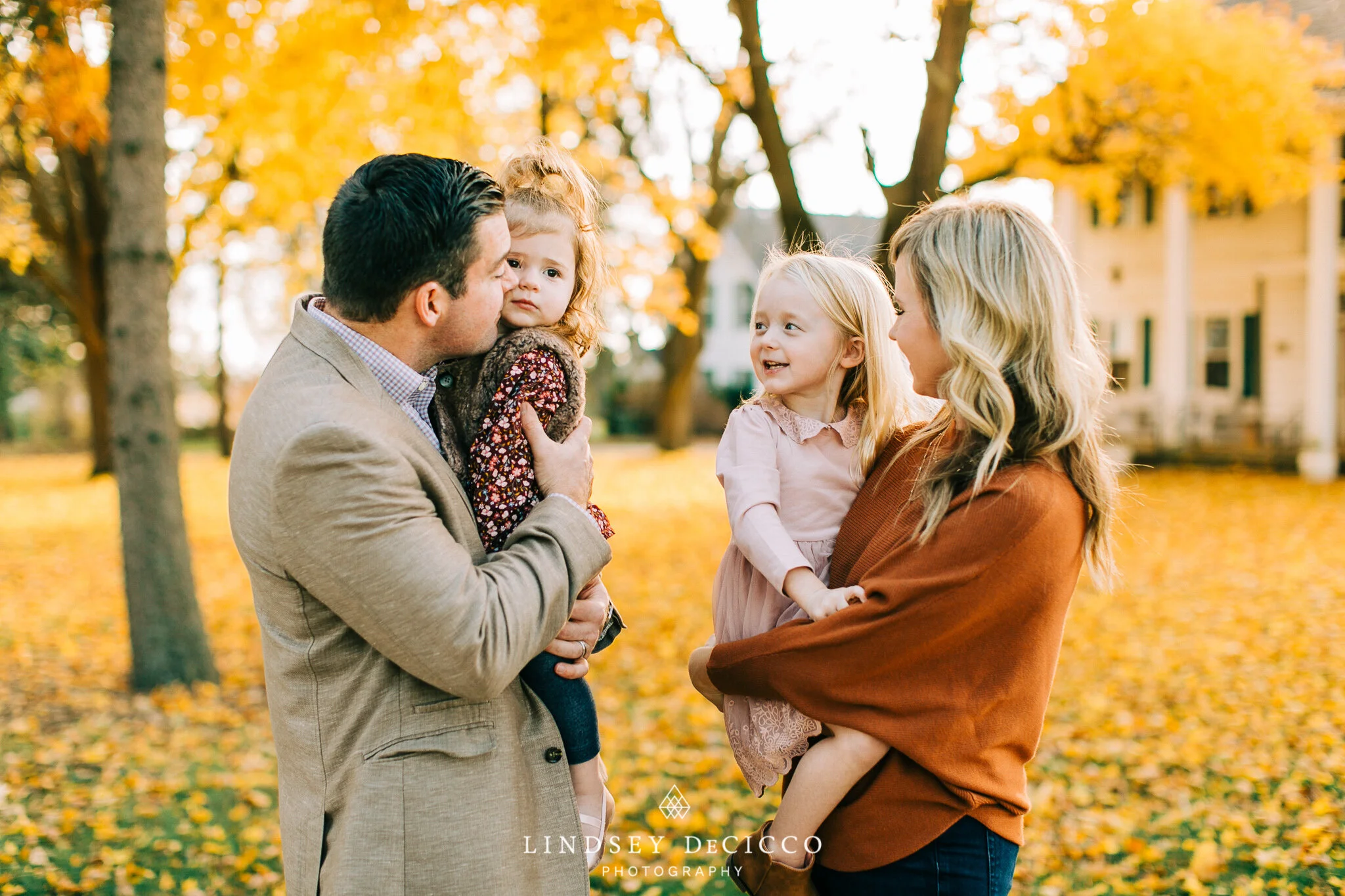 A joyful family of four stands together under vibrant golden fall trees, with the father kissing one daughter on the cheek while the mother holds their other daughter, all smiling during a warm autumn family photo session in Granger, Indiana.