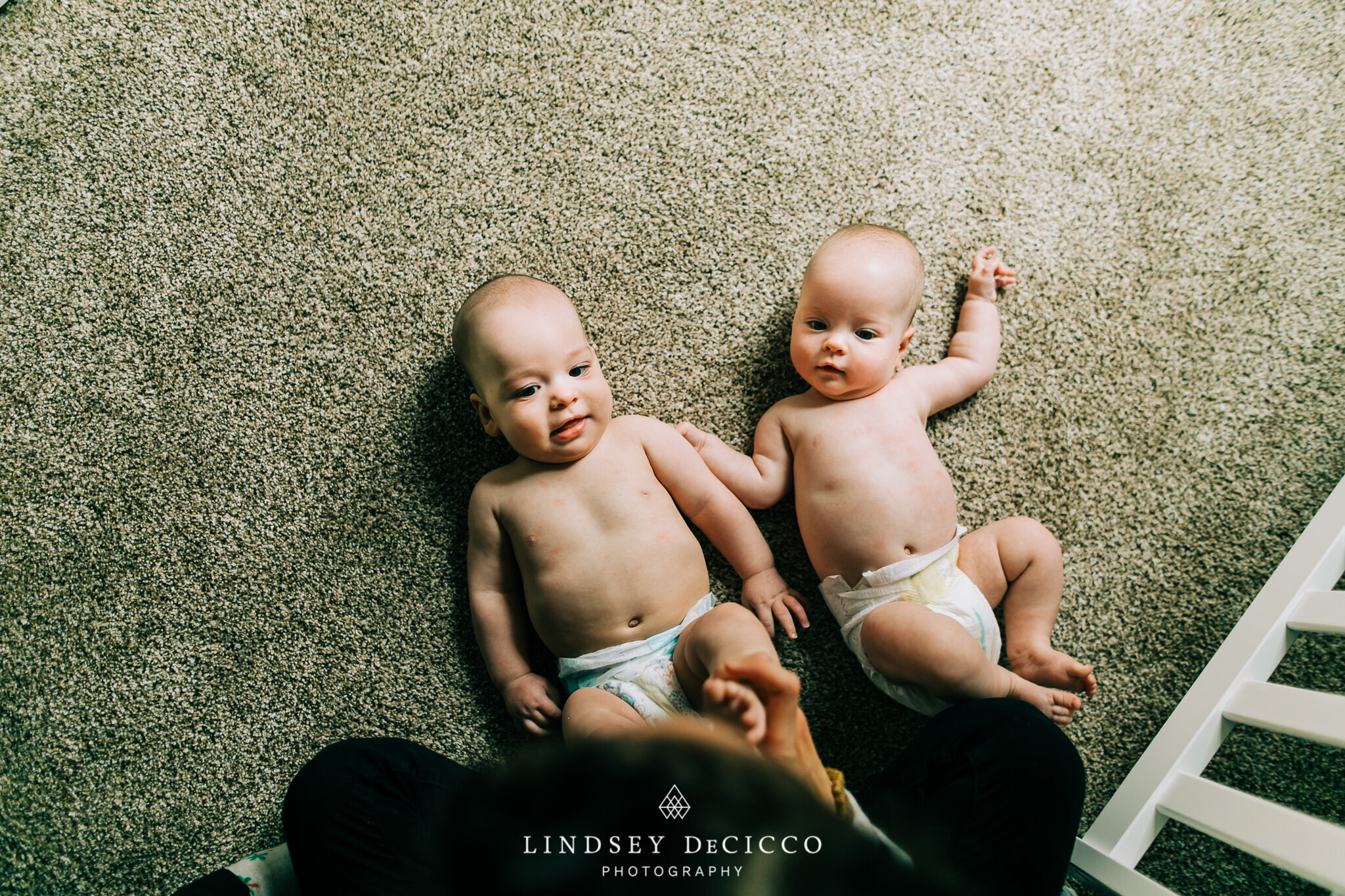 Two six-month-old twin babies lie side-by-side on a soft carpet, both wearing only diapers. One baby smiles softly while the other raises an arm, their little hands almost touching in a sweet, natural moment from their Grow-With-Me session.