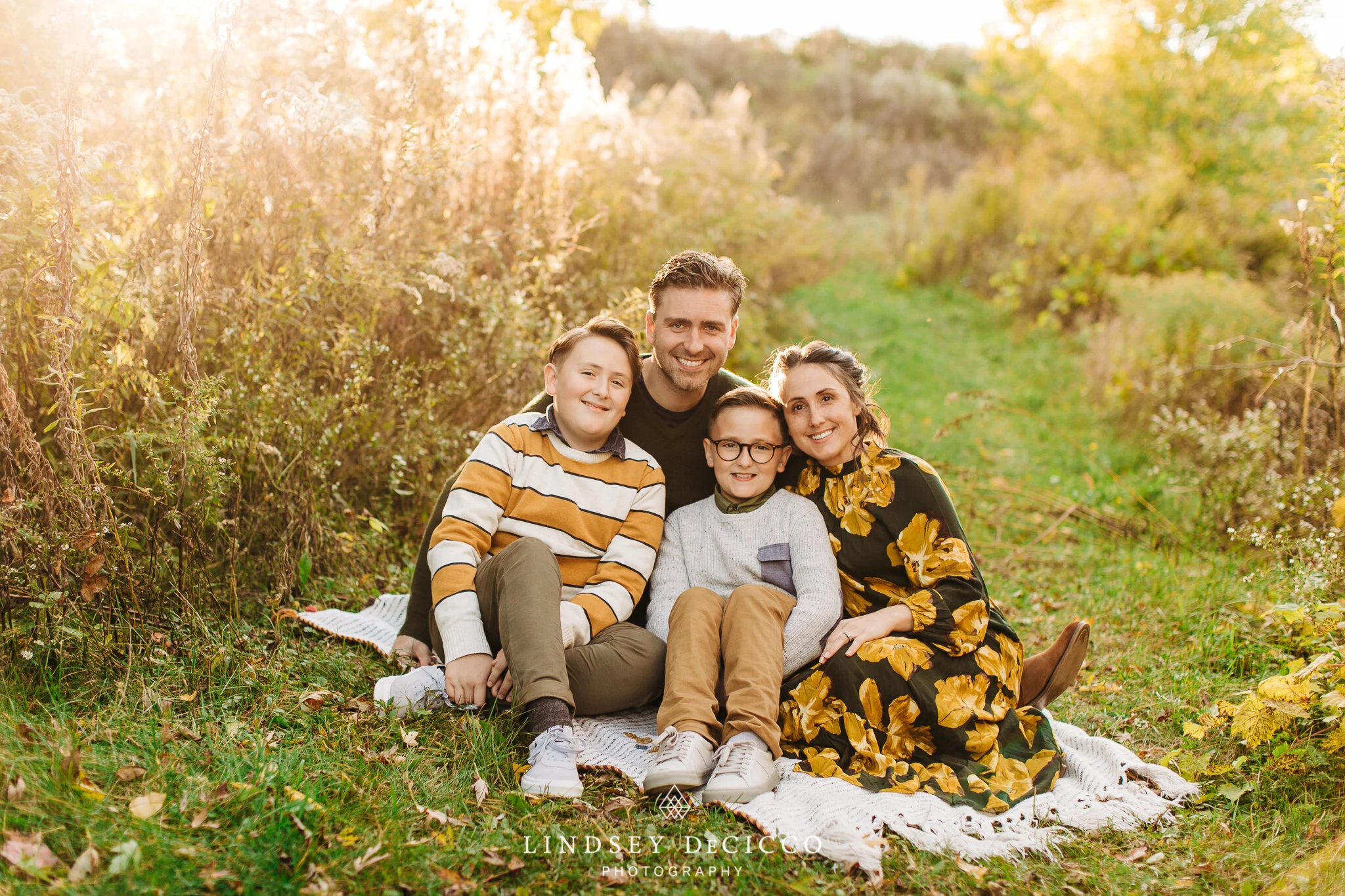 A family of four sits closely on a blanket in a sunlit field during golden hour. The parents smile warmly behind their two boys, who sit in front. Fall colors surround the scene, creating a soft and glowing atmosphere.