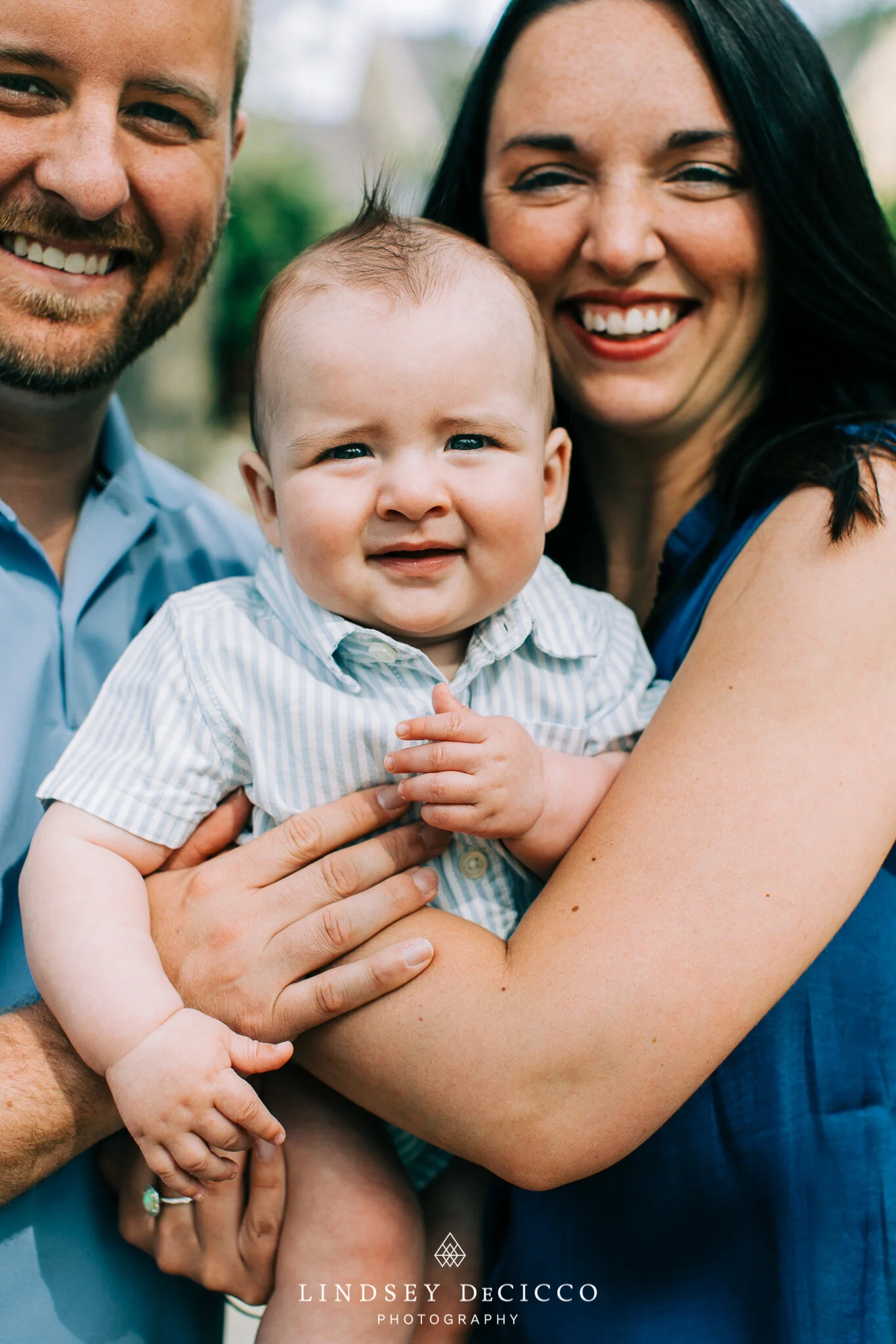 A baby boy is held between his smiling parents during an outdoor family photo session. The parents frame him closely, showing warm expressions and natural light highlighting the joyful moment.