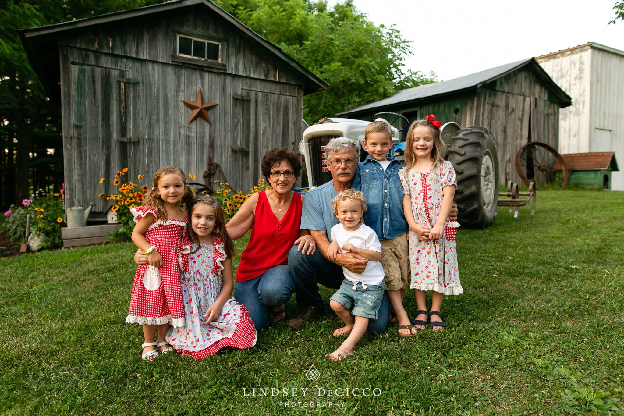 Grandma and Grandpa pose with their five young grandchildren in front of rustic barns and a vintage tractor at their Plymouth farmhouse, capturing a sweet summer moment.
