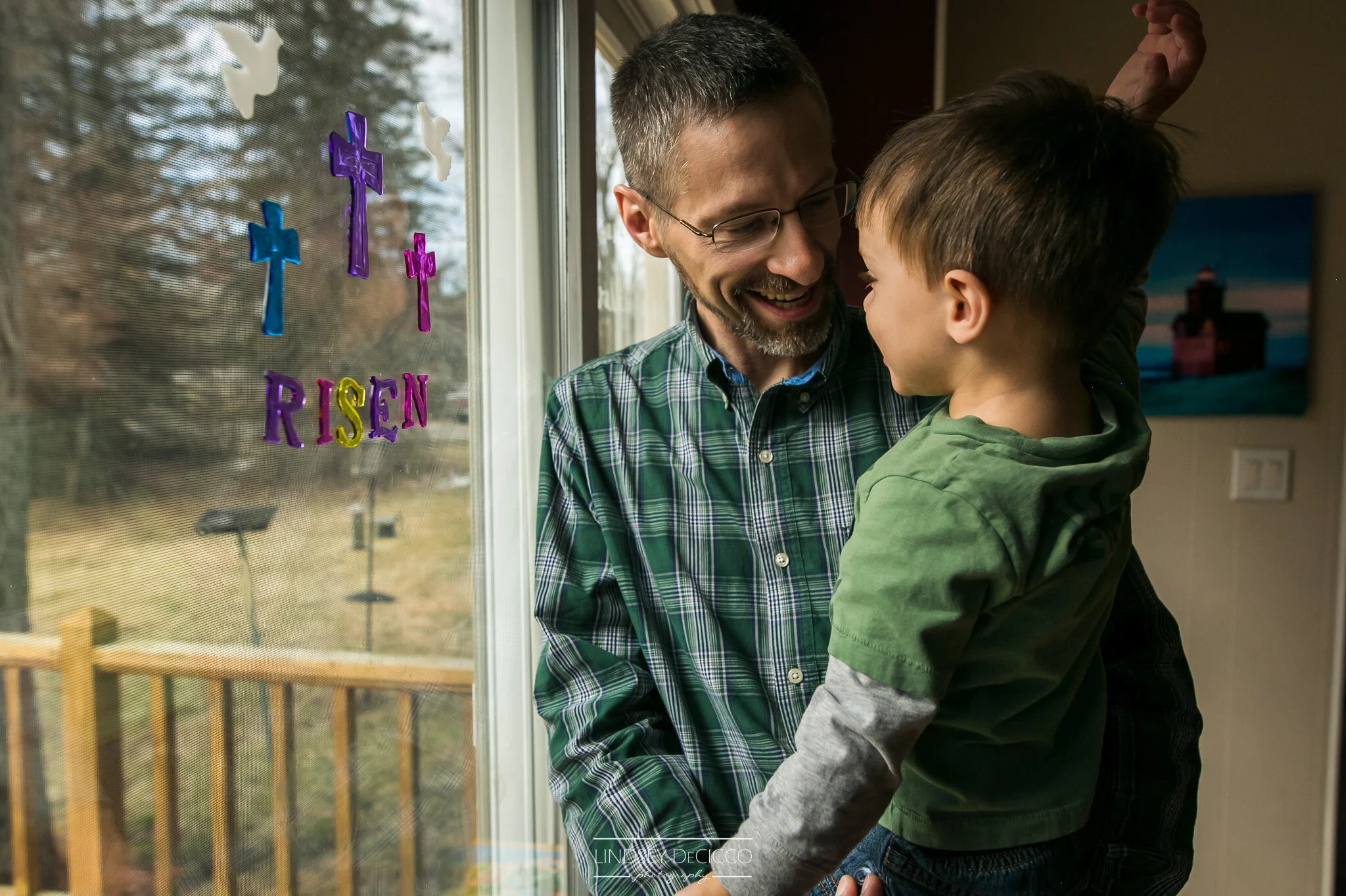A father smiles as he lifts his young son in front of a window decorated with colorful Easter-themed decals, both sharing a joyful moment together in natural light.