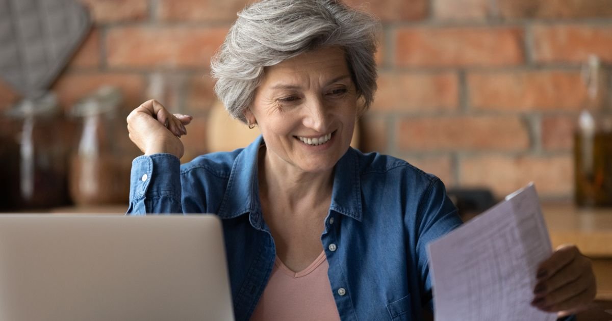 Happy mature woman reading financial documents on computer and paper