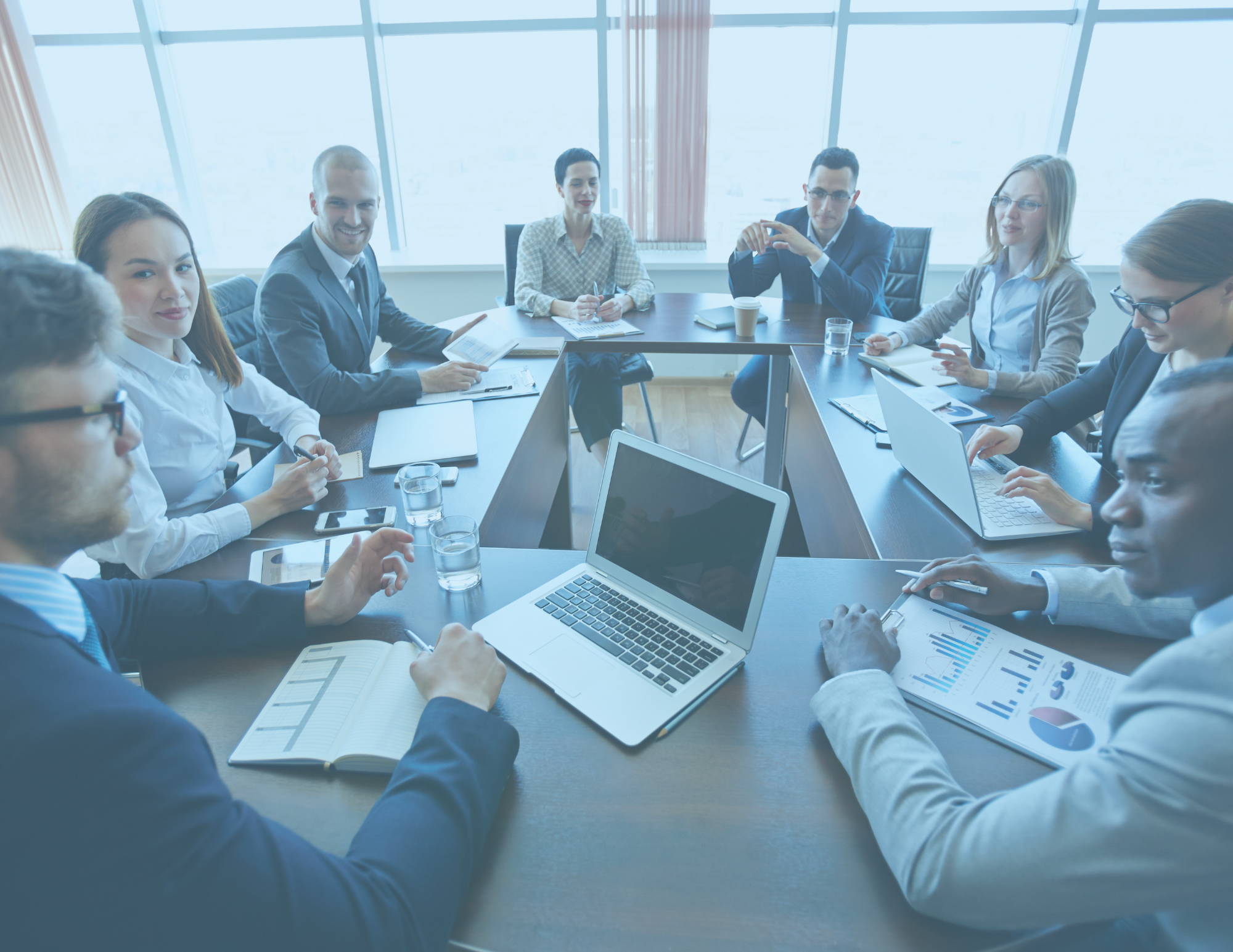 A group of eight professionals sitting around a conference table in a modern office during a meeting, with laptops, notebooks, and drinks.