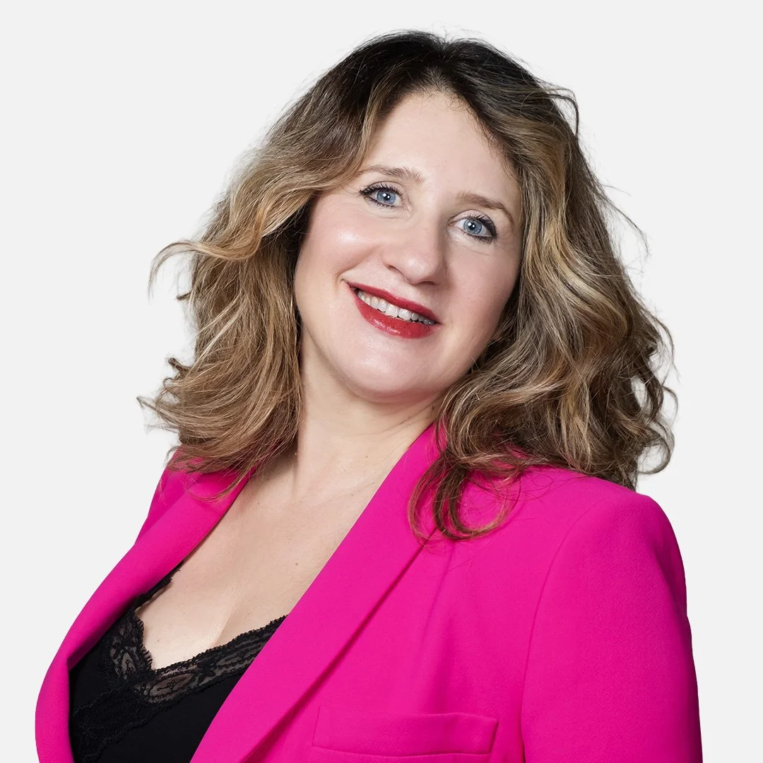 Donna con capelli biondo-scuro, indossa una giacca fucsia e una maglietta nera, sfondo bianco.