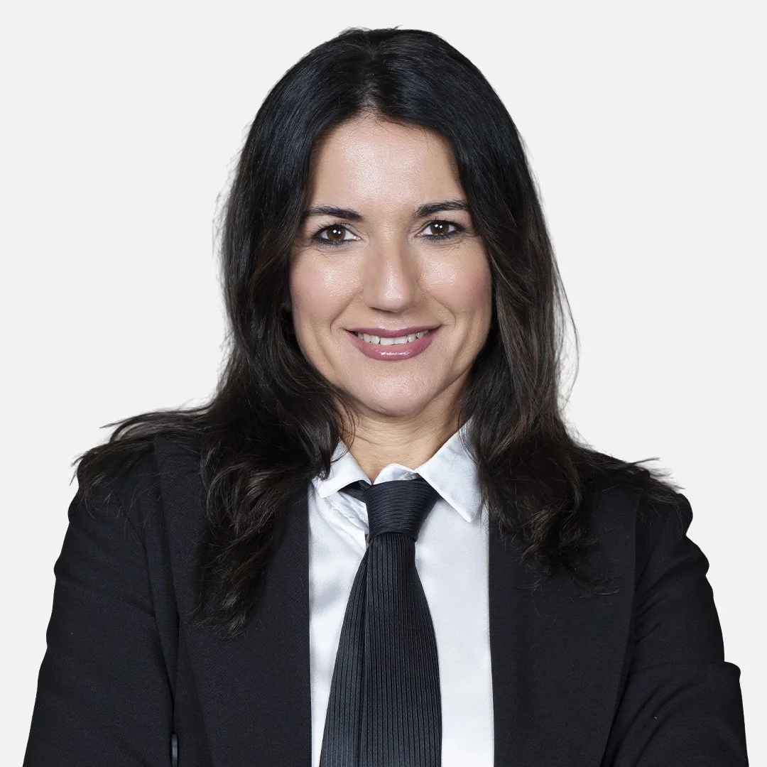 Donna con capelli scuri, indossa una giacca nera, una camicia bianca e una cravatta nera, sfondo bianco