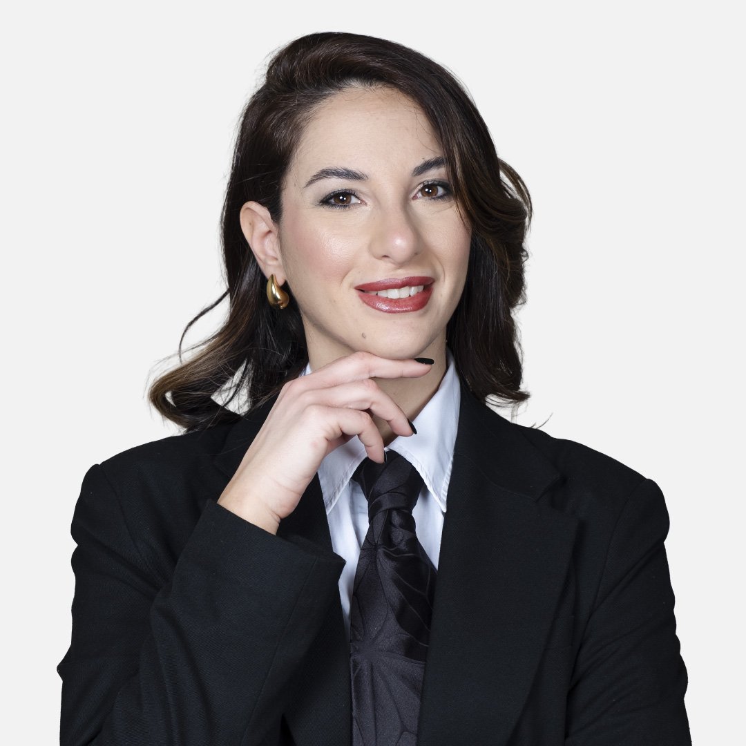 Donna con capelli scuri, indossa una giacca nera, una camicia bianca e una cravatta nera, sfondo bianco.