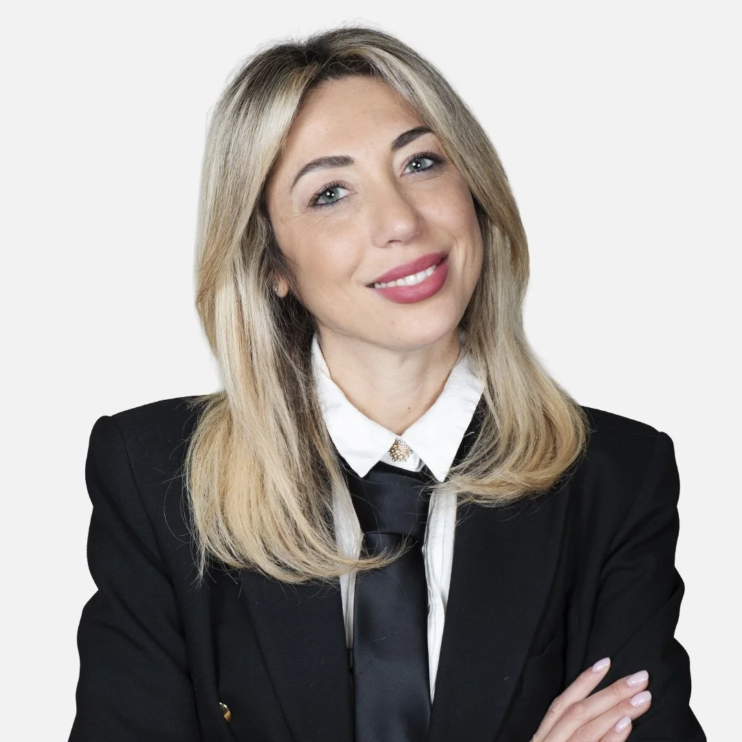 Donna con capelli biondi, indossa una giacca nera, una camicia bianca e una cravatta nera, sfondo bianco