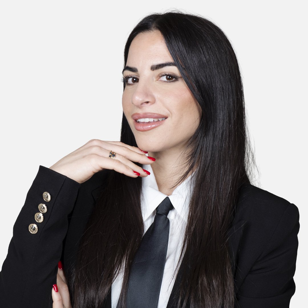 Donna con capelli lunghi e scuri, in abito formale, su sfondo bianco.