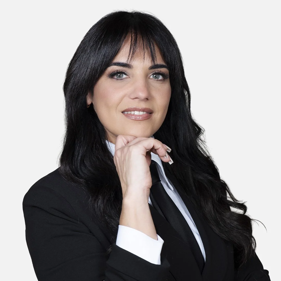 Donna con capelli scuri, indossa una giacca nera, una camicia bianca e una cravatta nera, sfondo bianco