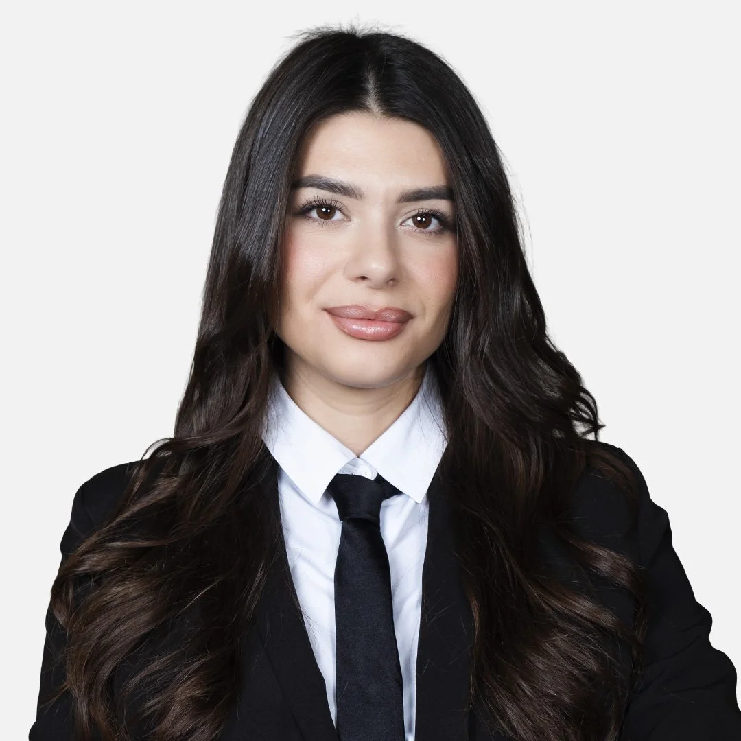Donna con capelli scuri, indossa una giacca nera, una camicia bianca e una cravatta nera, sfondo bianco.