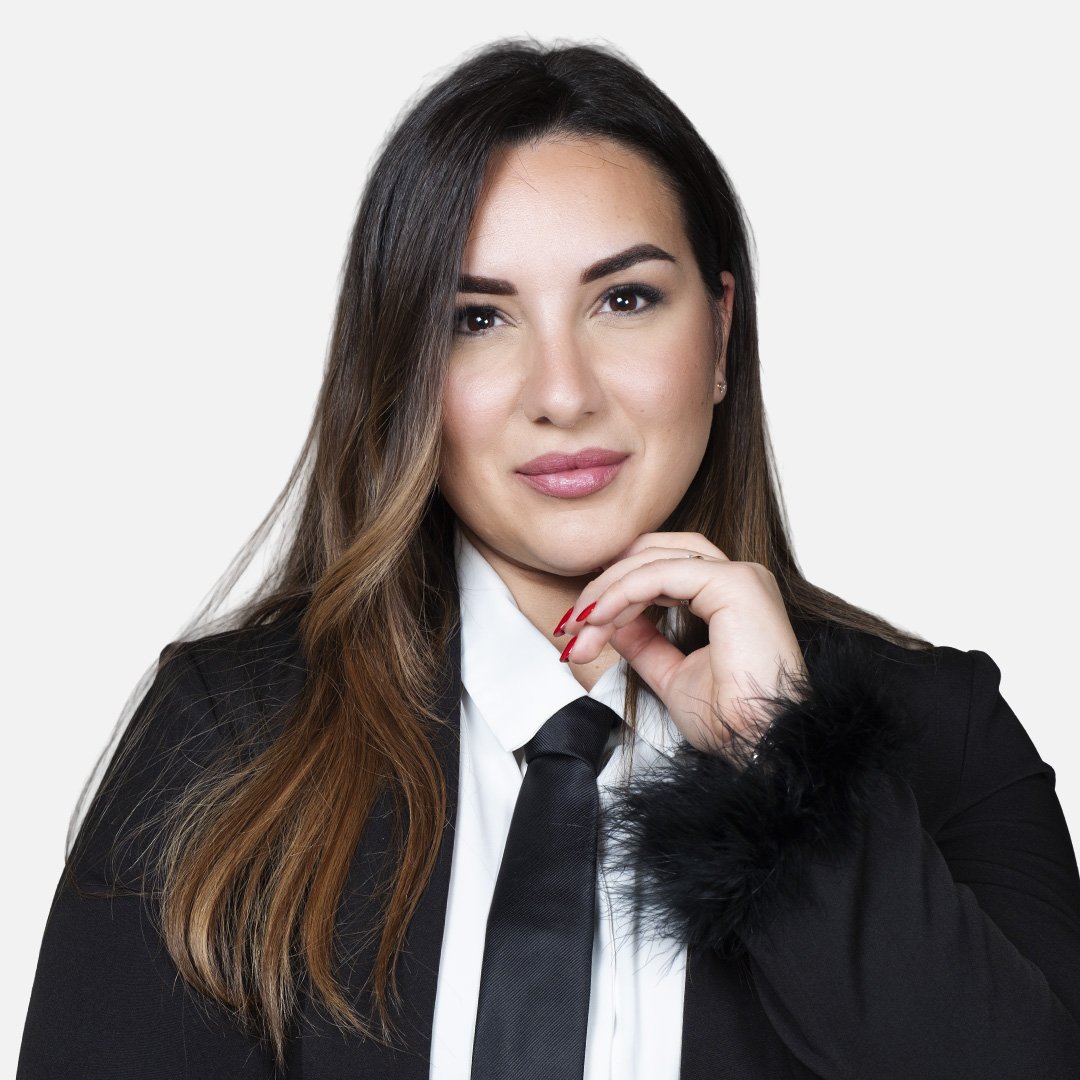 Donna con capelli castani, indossa una giacca nera e una camicia bianca con cravatta nera, sfondo bianco.