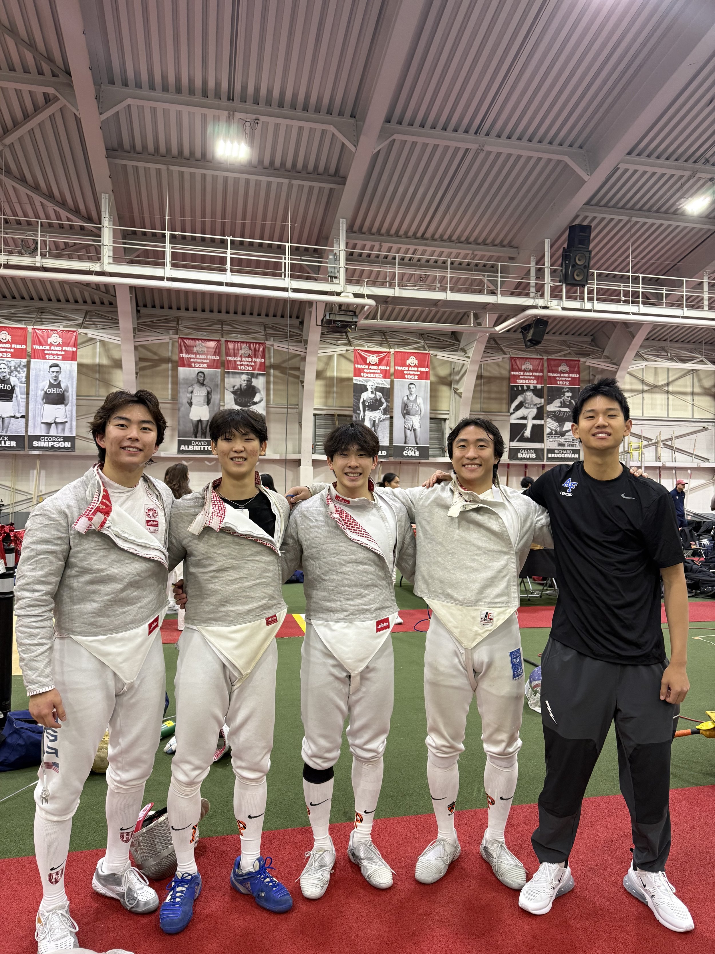 Group of five young male fencers standing together inside an indoor sports arena, smiling and posing for the photo. Four are dressed in fencing gear, and one is wearing a black sports shirt and pants.