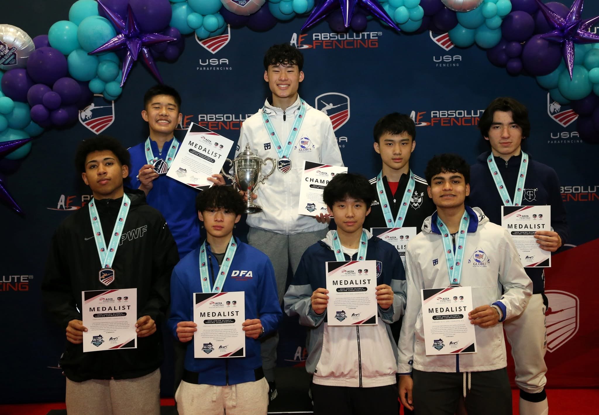 Young male athletes with medals and certificates at a fencing awards ceremony, standing on a podium in front of a decorated backdrop.