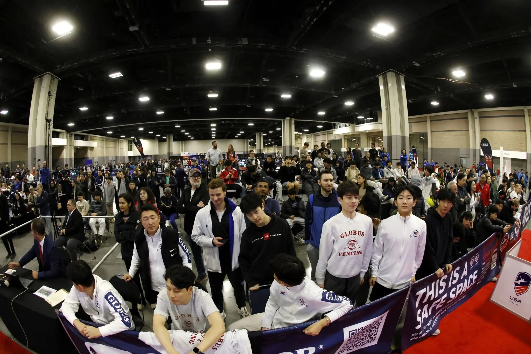Crowd of people attending an indoor event, with some seated at tables and others standing or walking, and a sign that says 'This is a Safe Space'.