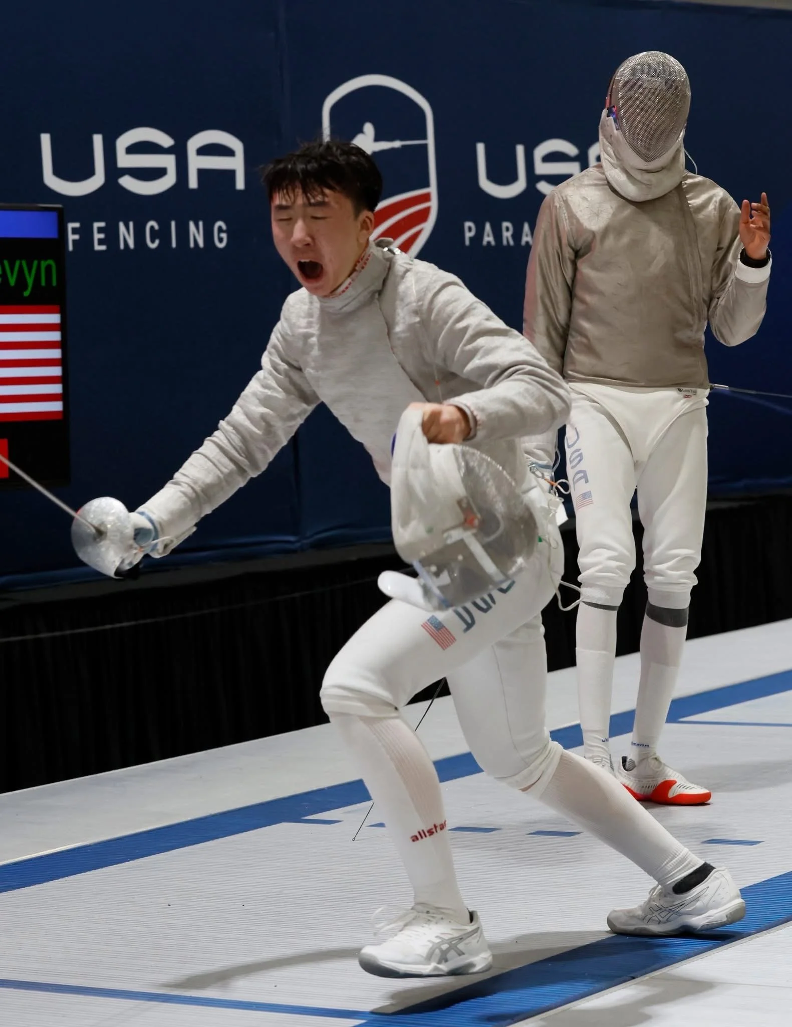 Two athletes in fencing gear celebrating during a match at a USA fencing event. One is kneeling with a clenched fist, and the other stands behind with a hand raised. Both wear white uniforms and masks, with a blue backdrop and the USA Fencing logo.