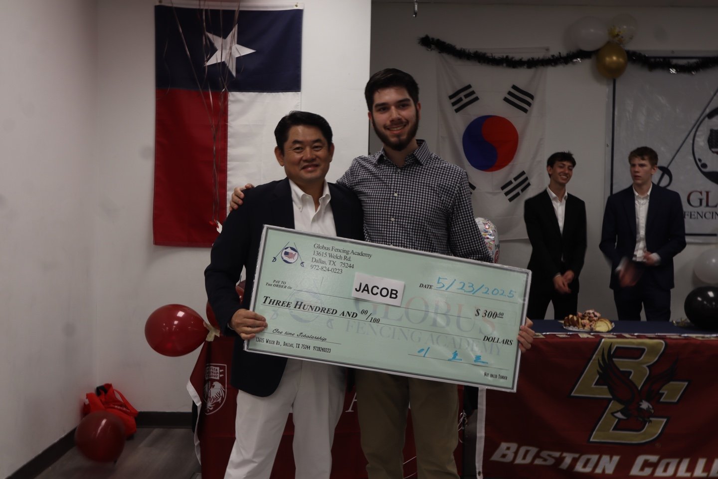 A young man named Jacob holding a large check for $300 at a ceremony. He is standing next to an older man. Behind them hangs a Texas flag and a South Korean flag. Four other young men in suits are in the background.