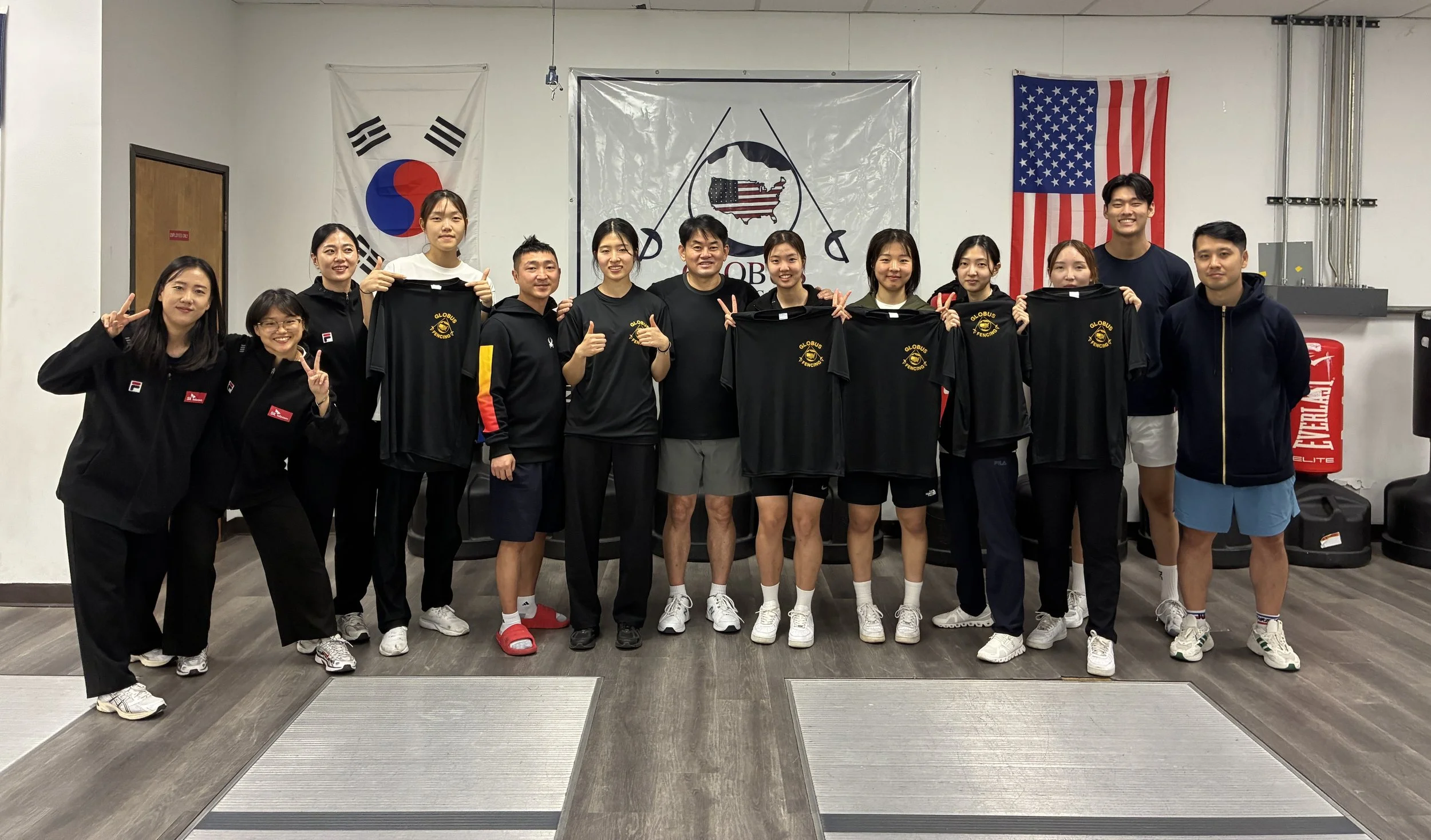 Group of people posing indoors in front of flags, including South Korea and the United States, holding black T-shirts with yellow logos, some making peace signs and thumbs-up gestures.