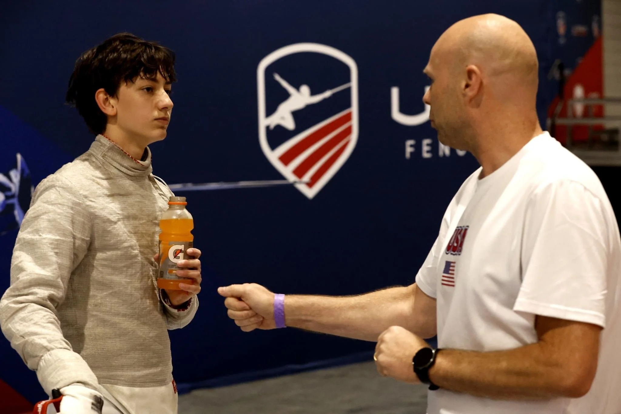 A young female fencer holding an orange Gatorade bottle, having a conversation with a bald male coach or official, in front of a US Fencing logo.