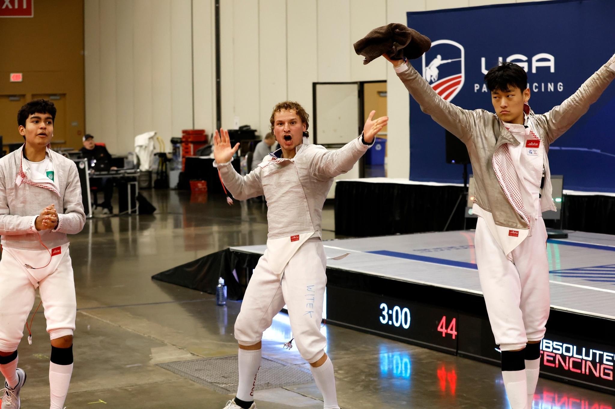 Three young fencers in a sports hall, dressed in fencing gear, celebrating or reacting during a match at the U.S. Fencing League event with a digital scoreboard and fencing platform in the background.
