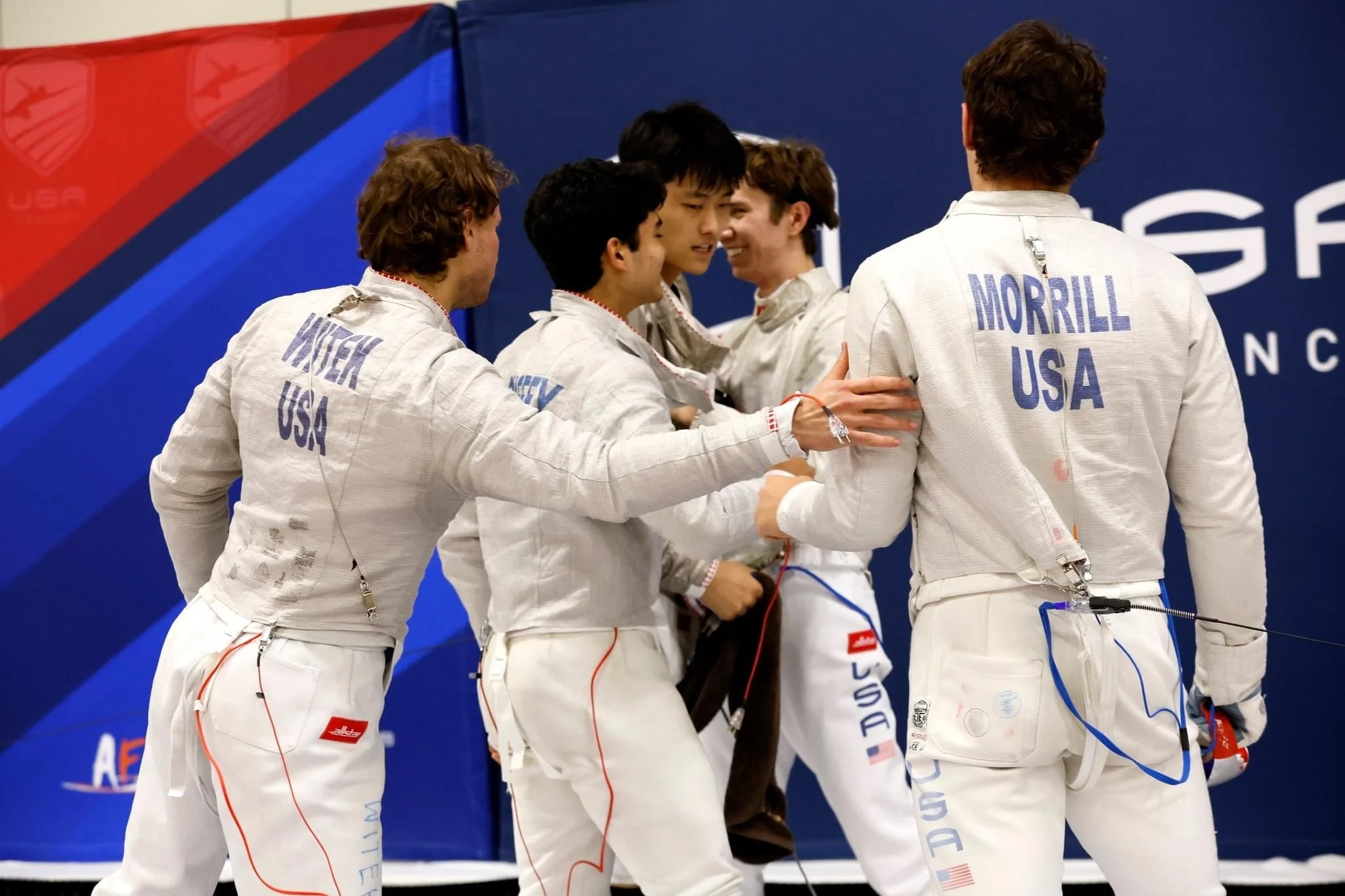 Fencing team from the USA celebrating after a match, wearing white uniforms with their country and last names on the back, on a blue background with fencing-related branding.