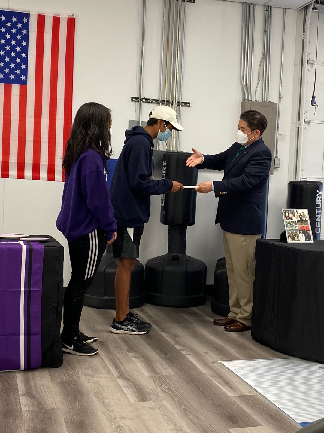 A man in a suit and face mask is handing a certificate to a young man in a hoodie, shorts, and face mask, with a young woman in casual clothes watching. There is an American flag on the wall behind them.