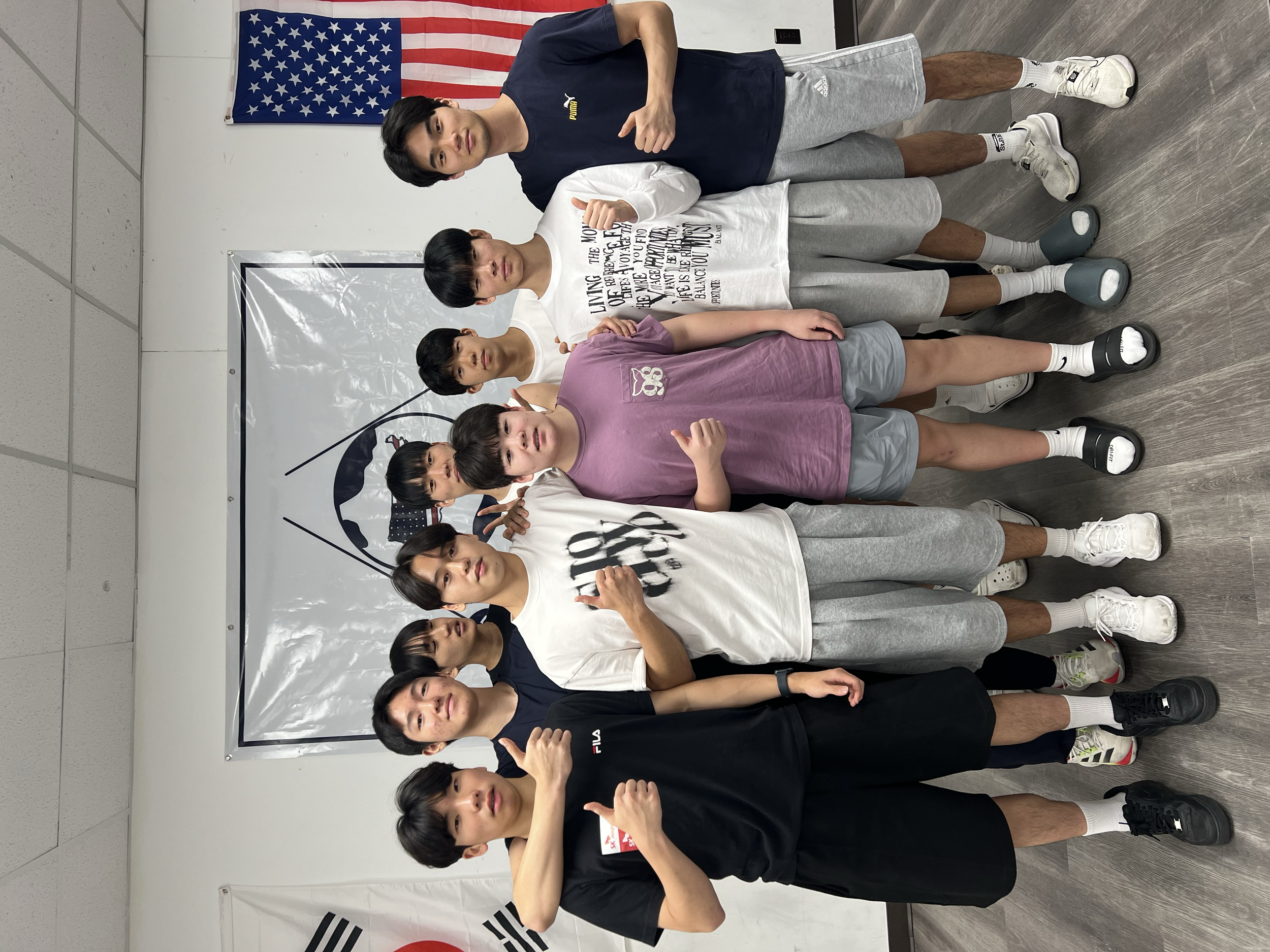 Group of young athletes posing indoors in front of a whiteboard with flags, wearing sportswear and making various hand gestures.