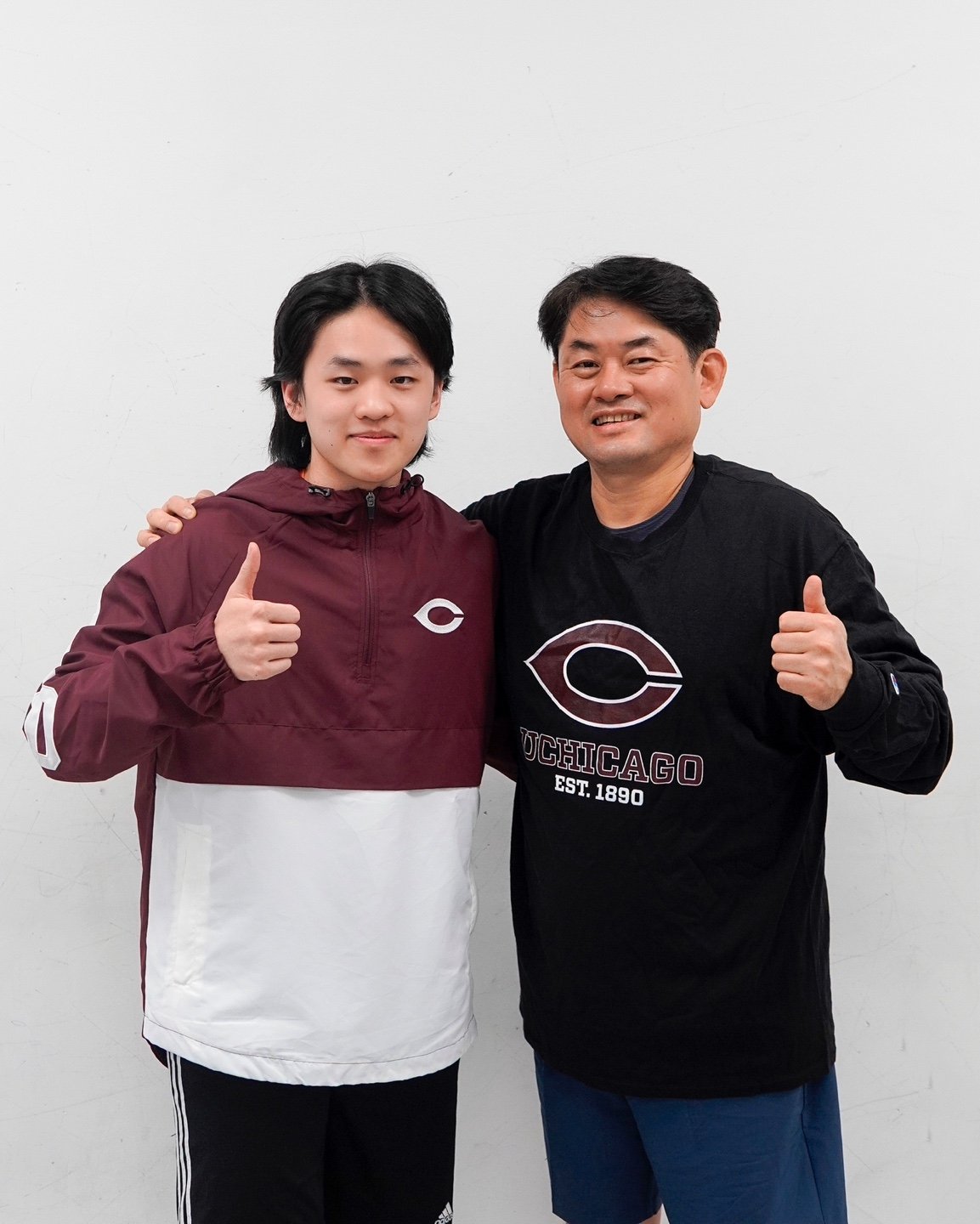 Two men pose for a photo against a plain white wall, wearing Chicago sports apparel and giving thumbs-up gestures.