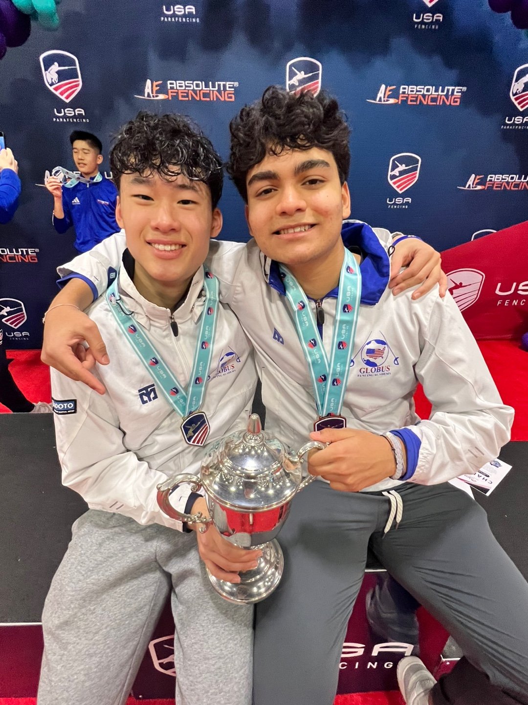 Two young male athletes smiling and sitting together, each wearing white jackets with blue accents and medals around their necks, holding a large silver trophy in front of a backdrop with logos, including 'USA Fencing' and 'Absolute Fencing'.
