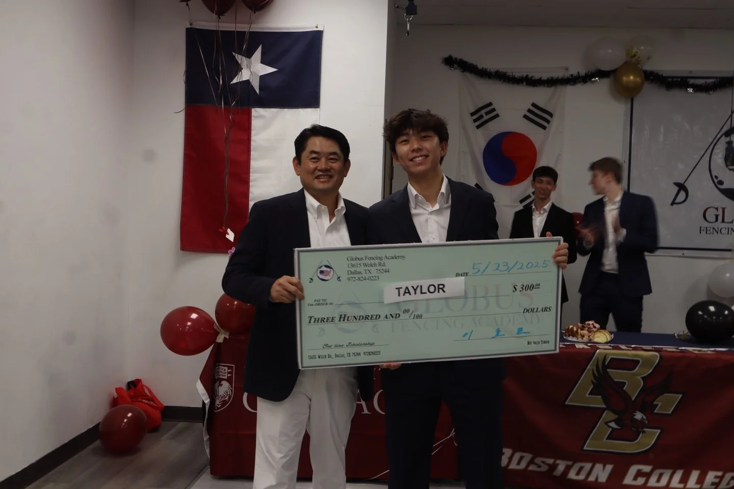 Two young men in suits holding a large check with a man on the left and a young man on the right. Behind them, there are three men clapping, a Texas flag, and a South Korean flag, and decorations like balloons and a banner for a fencing academy.