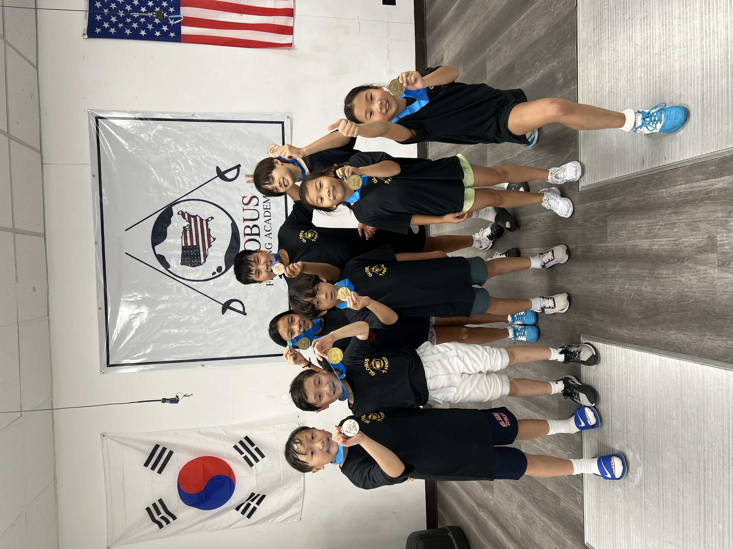 Group of children in sportswear holding medals and posing in a gym, with US and South Korean flags on the wall behind them.