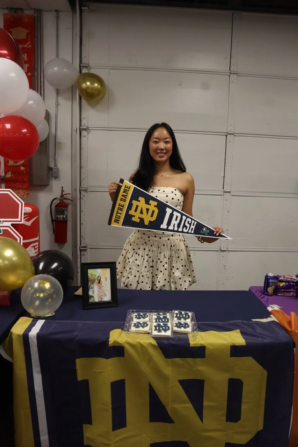 A young woman standing behind a table decorated with Notre Dame logos, holding a Notre Dame pennant, celebrating at a party with balloons and cookies, in a garage or basement setting.