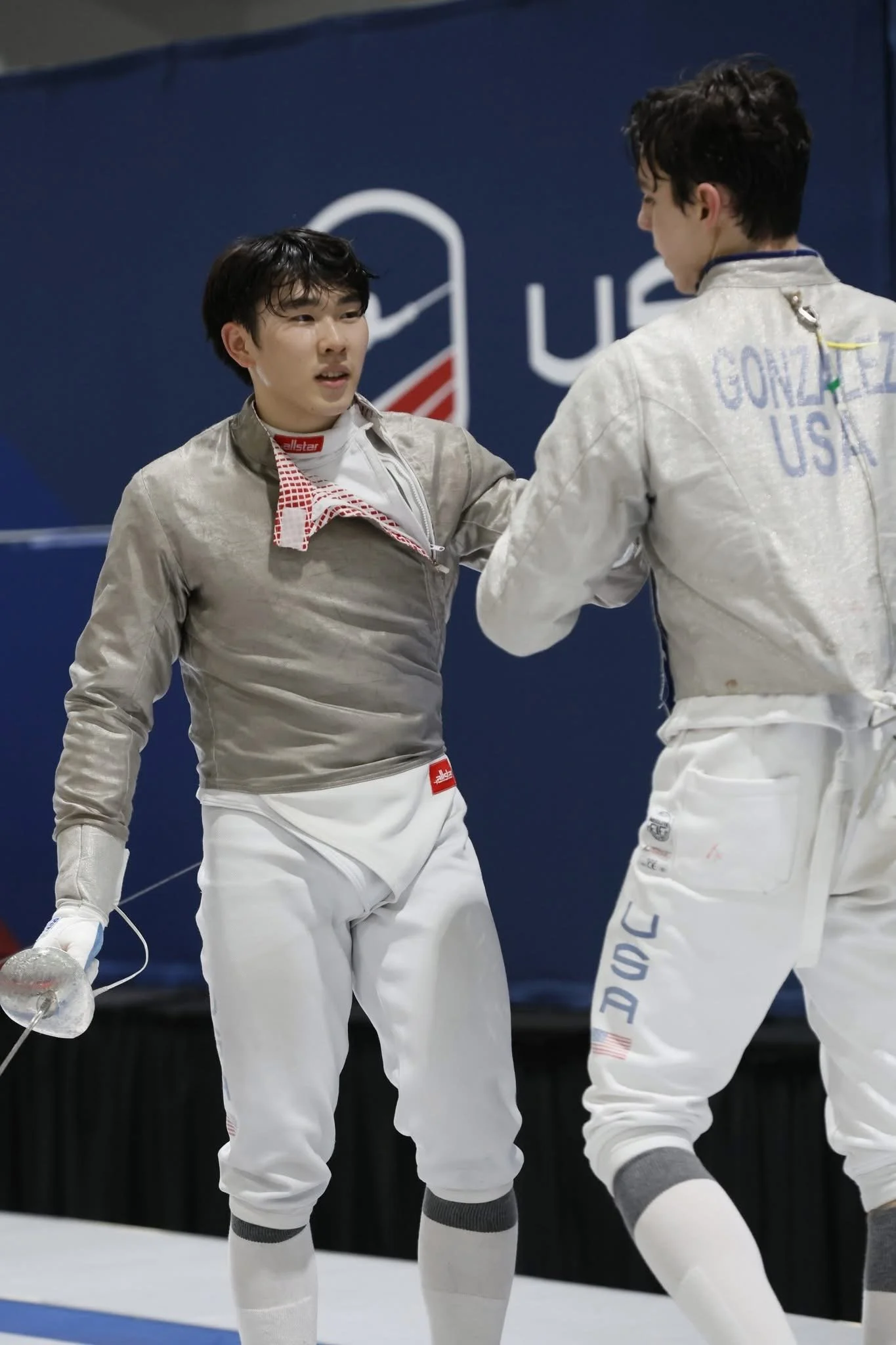 Two male Olympic foil fencers in fencing gear, engaging in a match, with one holding a foil and the other placing his hand on his opponent's shoulder.