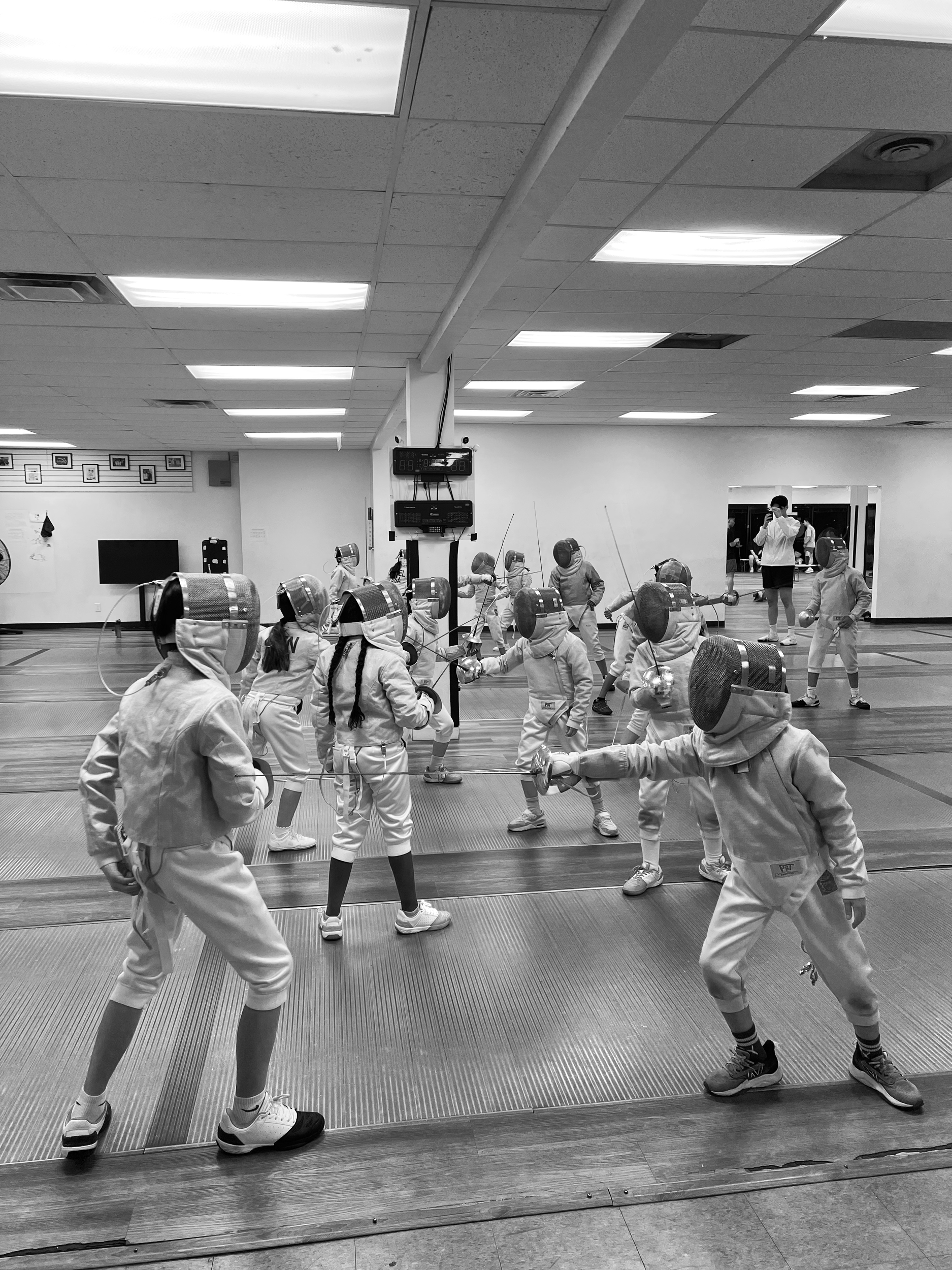 Children practicing fencing in a school gym, all wearing protective gear including masks and uniforms, with a coach observing in the background.