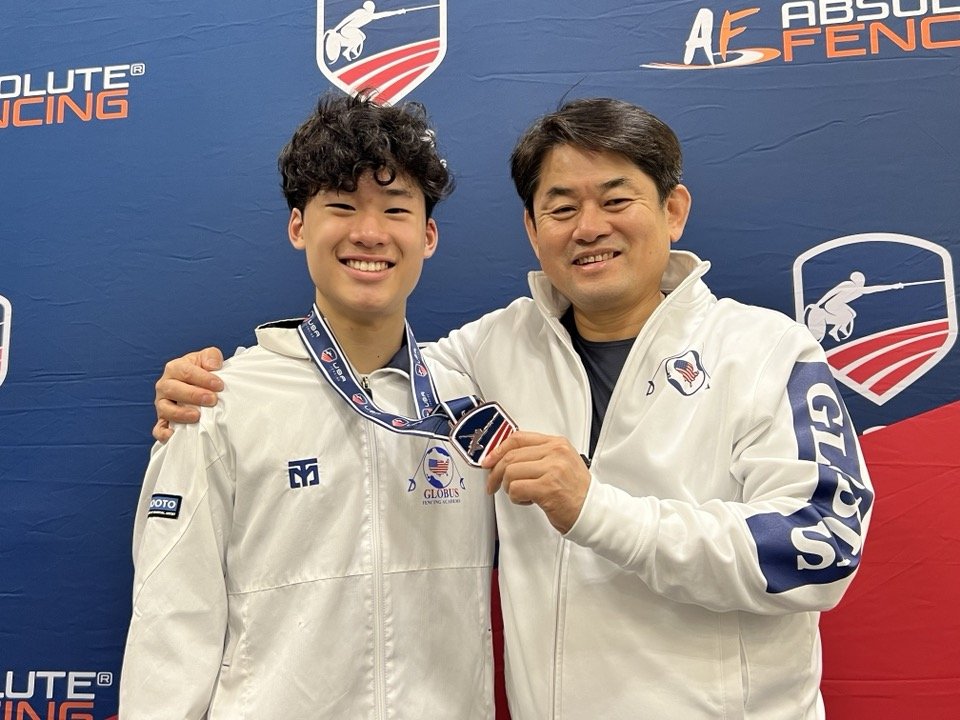 A young man and an older man are smiling and posing for a photo at an indoor fencing event. The young man is wearing a white fencing jacket and a medal around his neck, while the older man is also dressed in a white jacket. They are standing in front