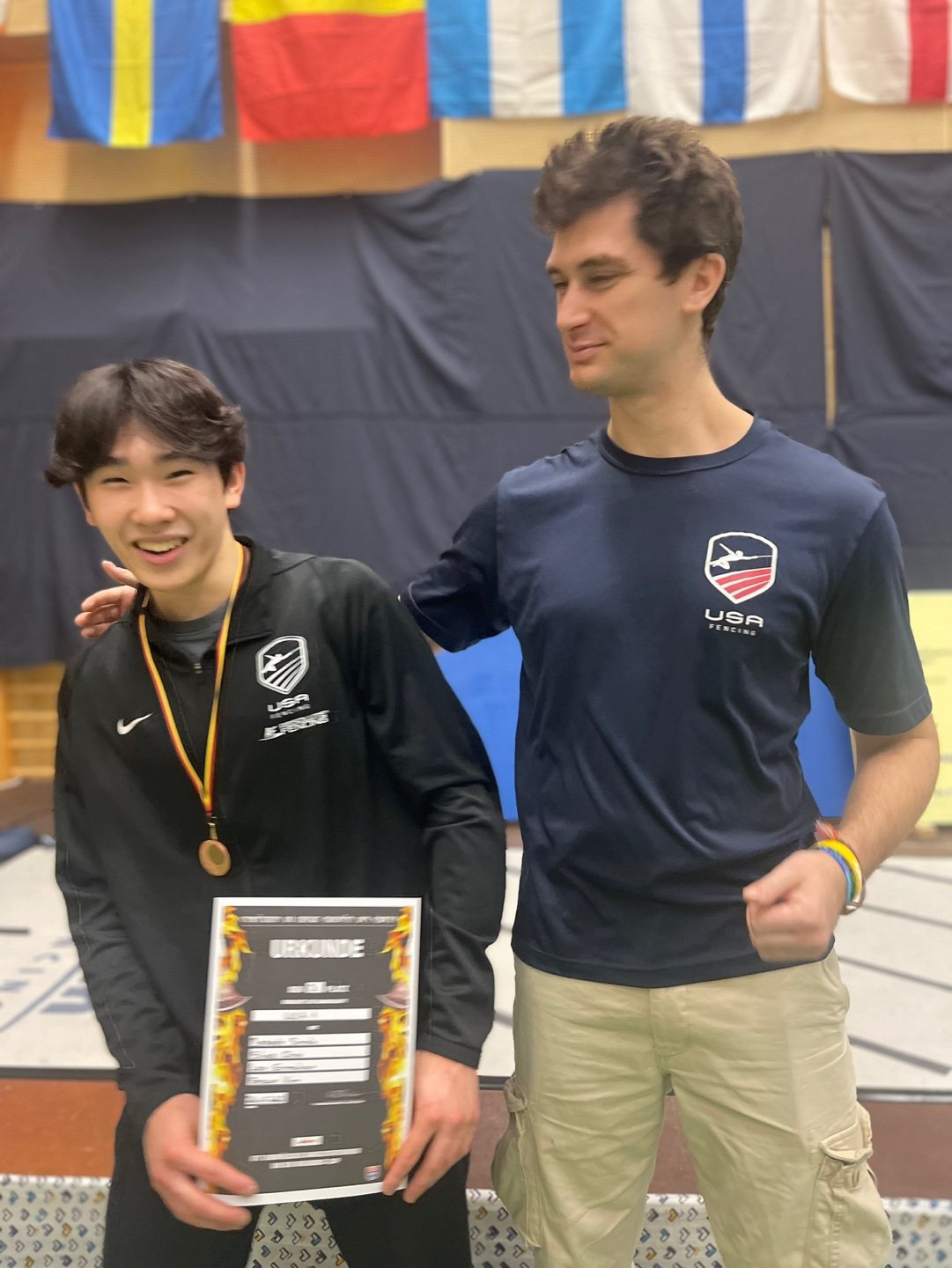 Two male athletes at an awards ceremony, one young with a medal and certificate, both wearing USA fencing shirts, with international flags hanging in the background.