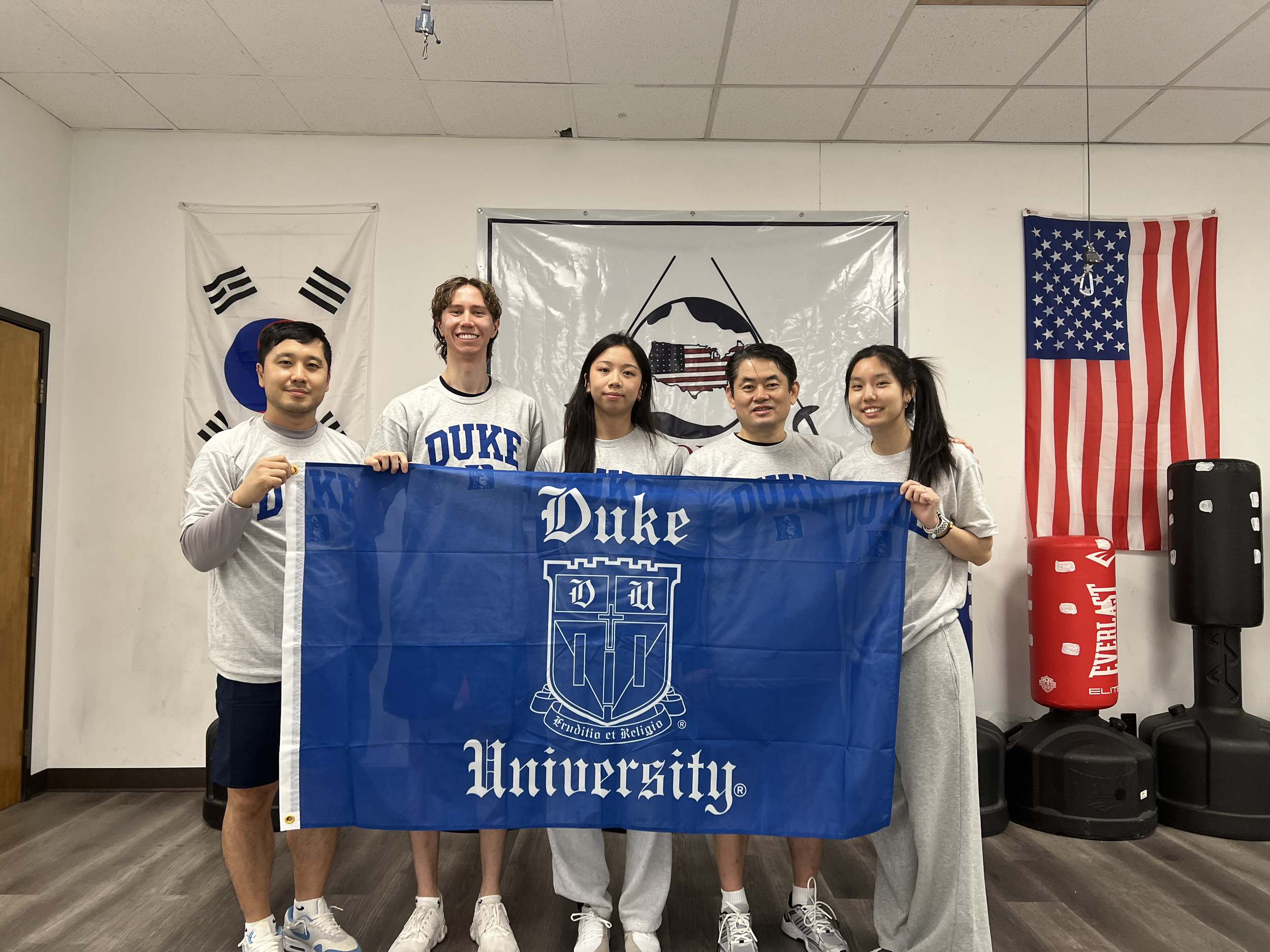 Group of six people holding a Duke University flag, standing in front of American, South Korean, and other flags in a room with boxing equipment.